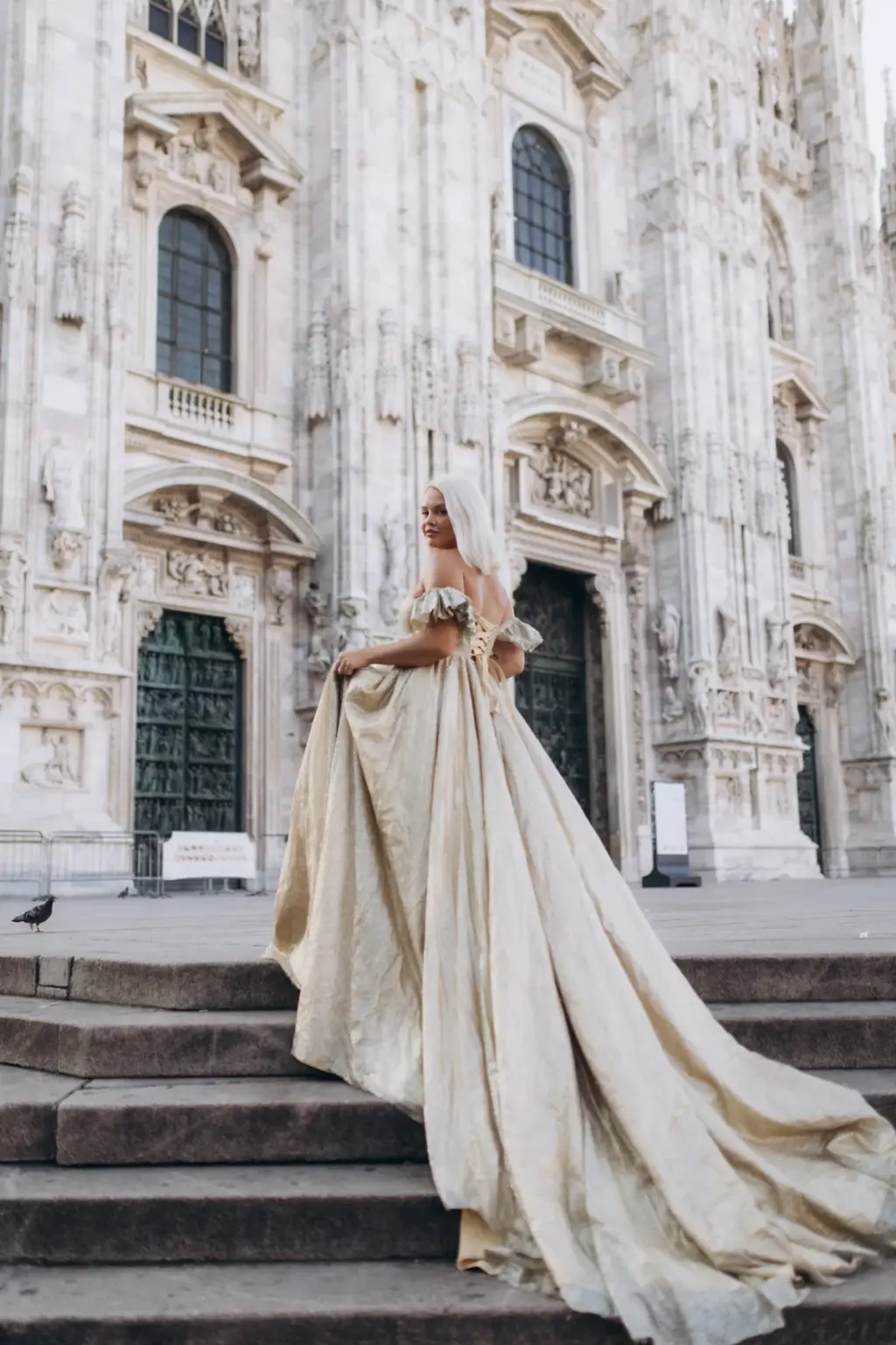 A woman in a long champagne flying dress stands on the Duomo steps, the train flowing down the stone stairs below the carved doors.