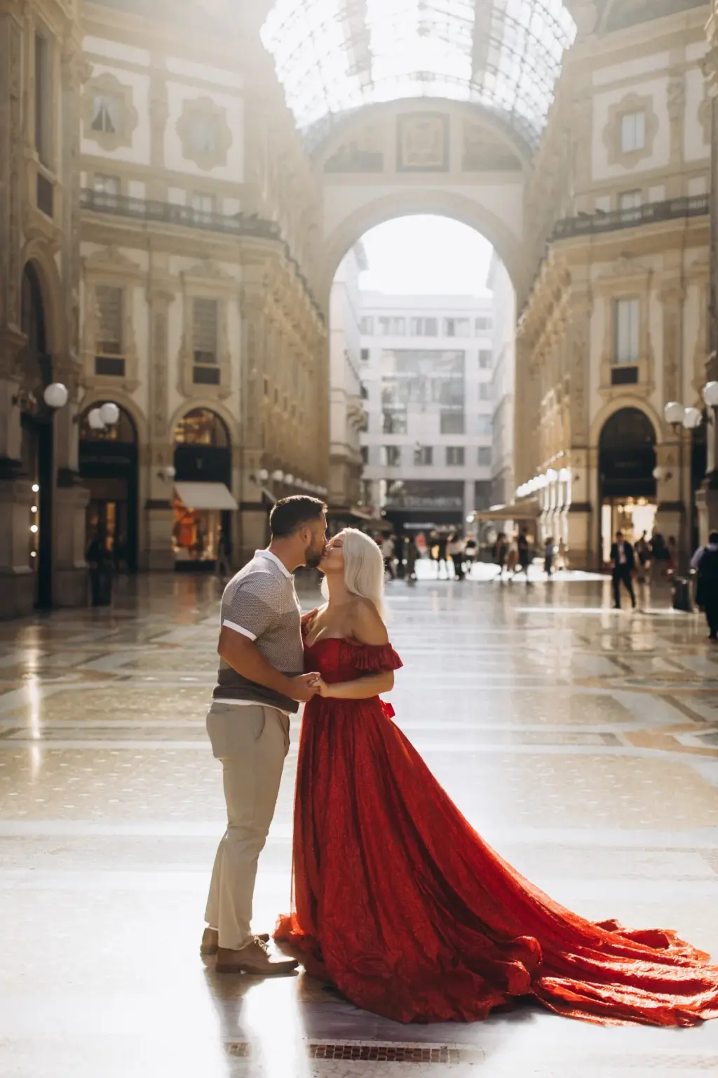 A man and a woman in a long red gown kiss on the gleaming floor of the Galleria, the glass roof and arched entrance behind them.