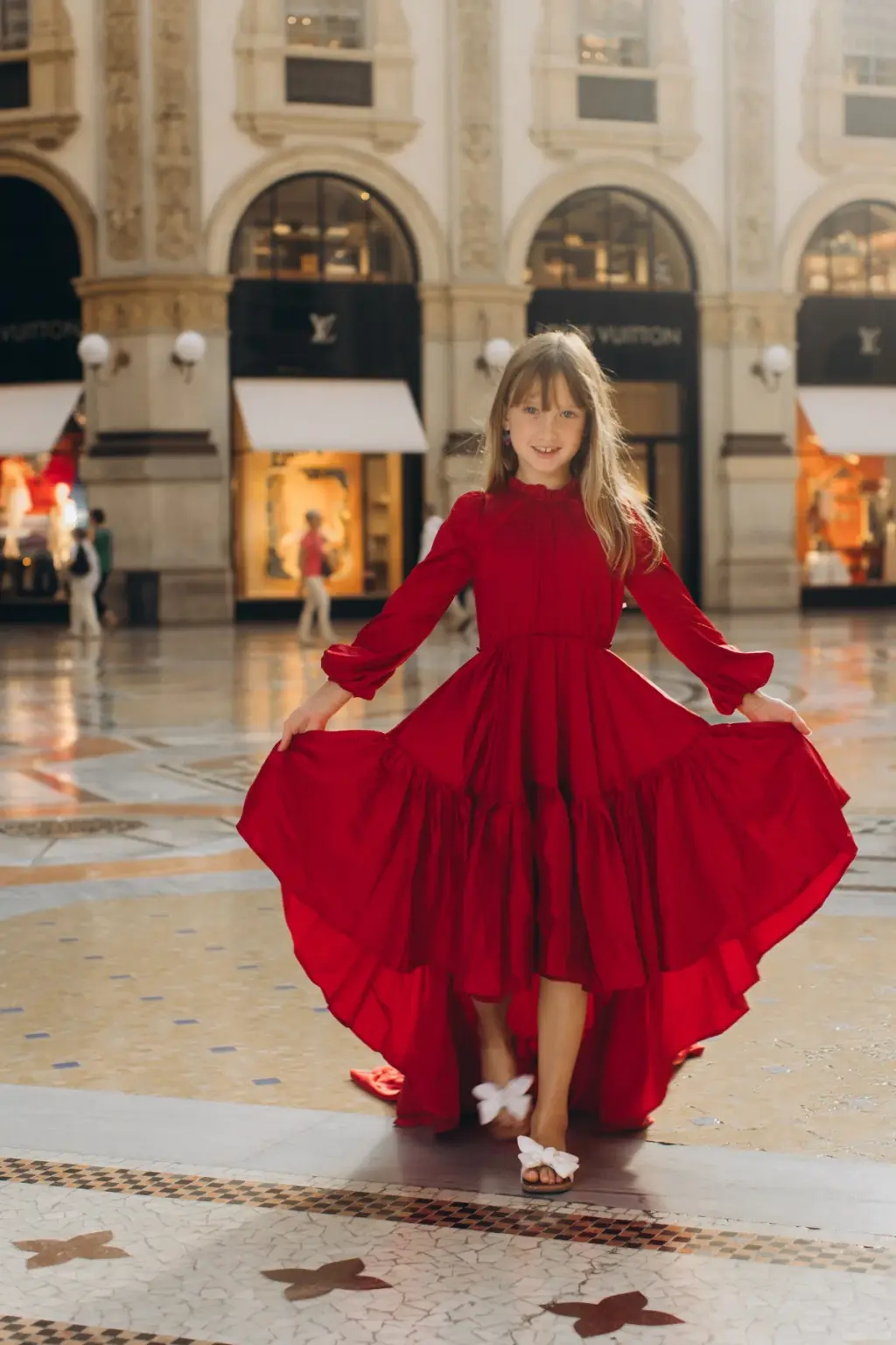 A little girl in a red high-low dress and white sandals holds out her skirt and smiles in the centre of the Galleria.