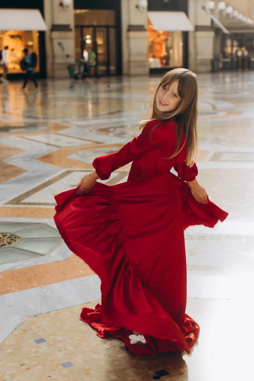 A young girl with long hair spins in a red tiered dress on the marble mosaic floor of the Galleria, smiling over her shoulder.