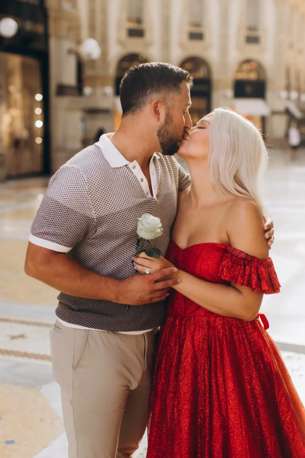 A bearded man holding a white rose kisses a blonde woman in a red off-the-shoulder gown inside the Galleria Vittorio Emanuele II.
