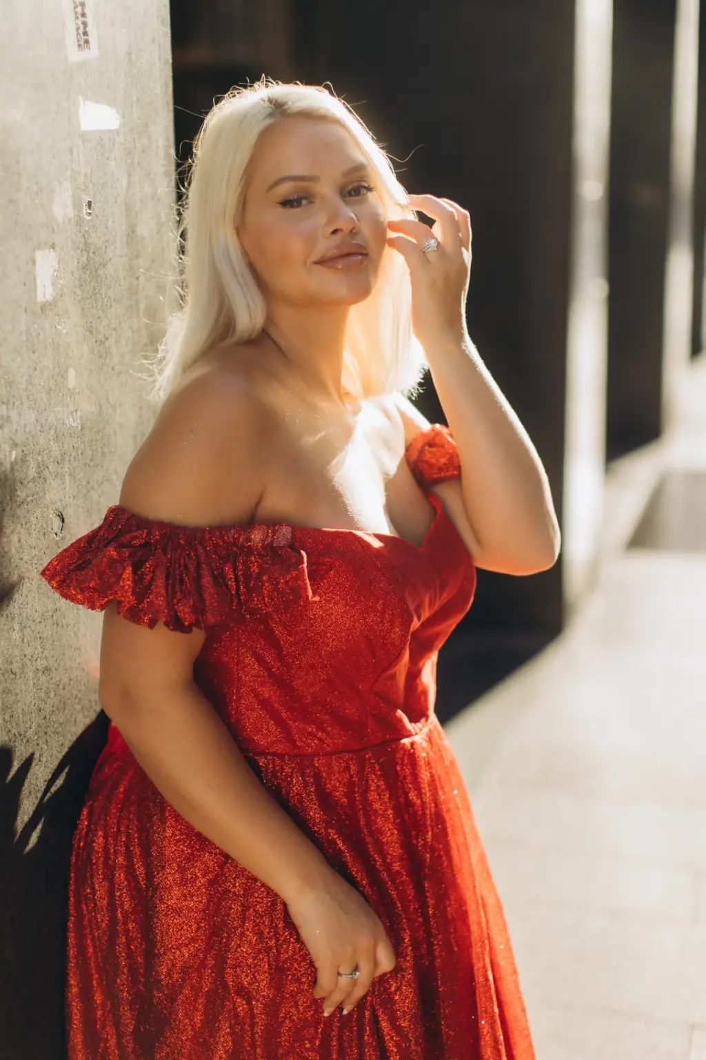 A blonde woman in a red off-the-shoulder dress leans on a stone pillar in warm light, fingers brushing her hair in a Milan arcade.