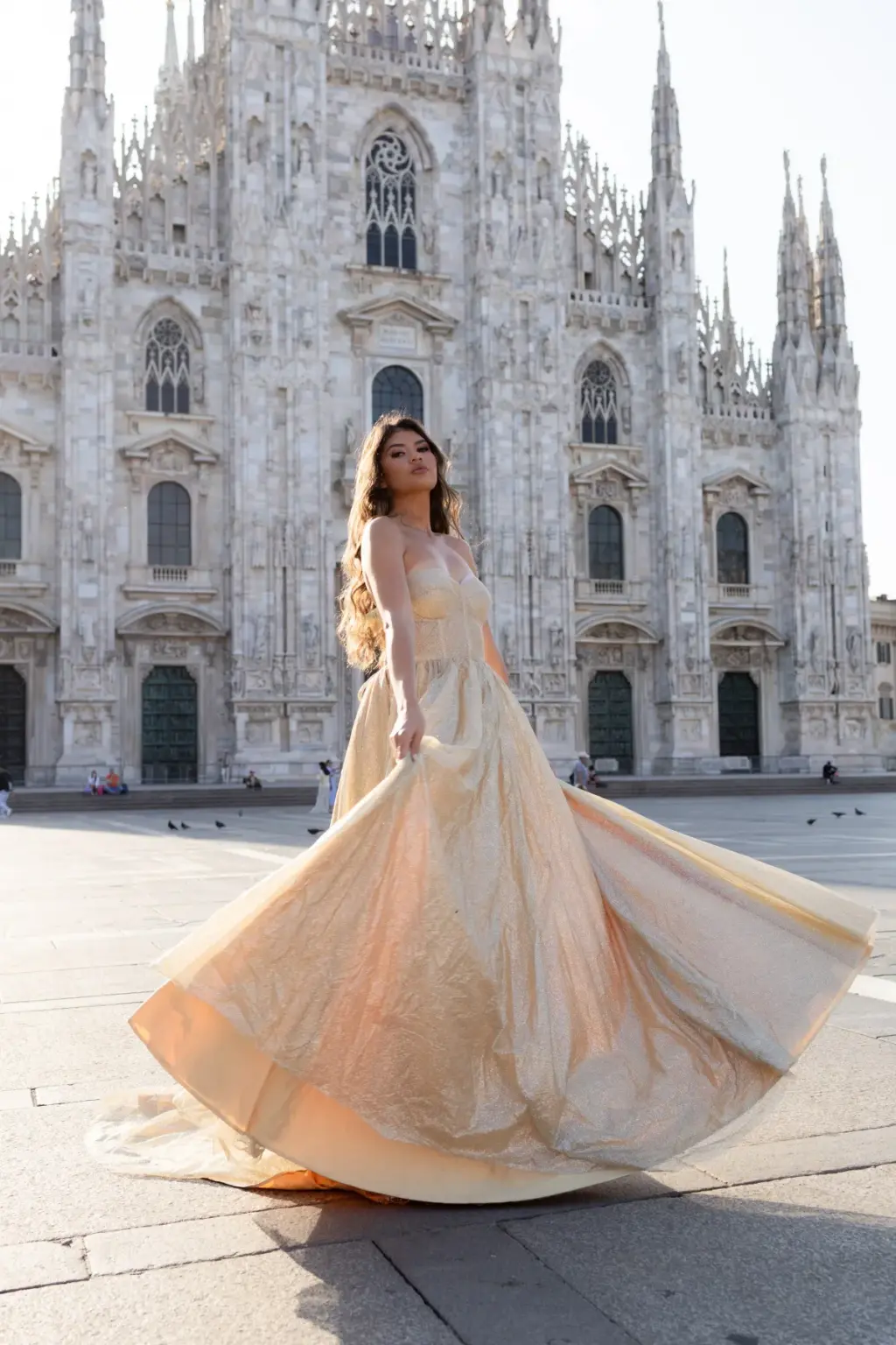 A woman sweeps the long skirt of her shimmering gold gown into a wide swirl on the paving stones before the Duomo di Milano.