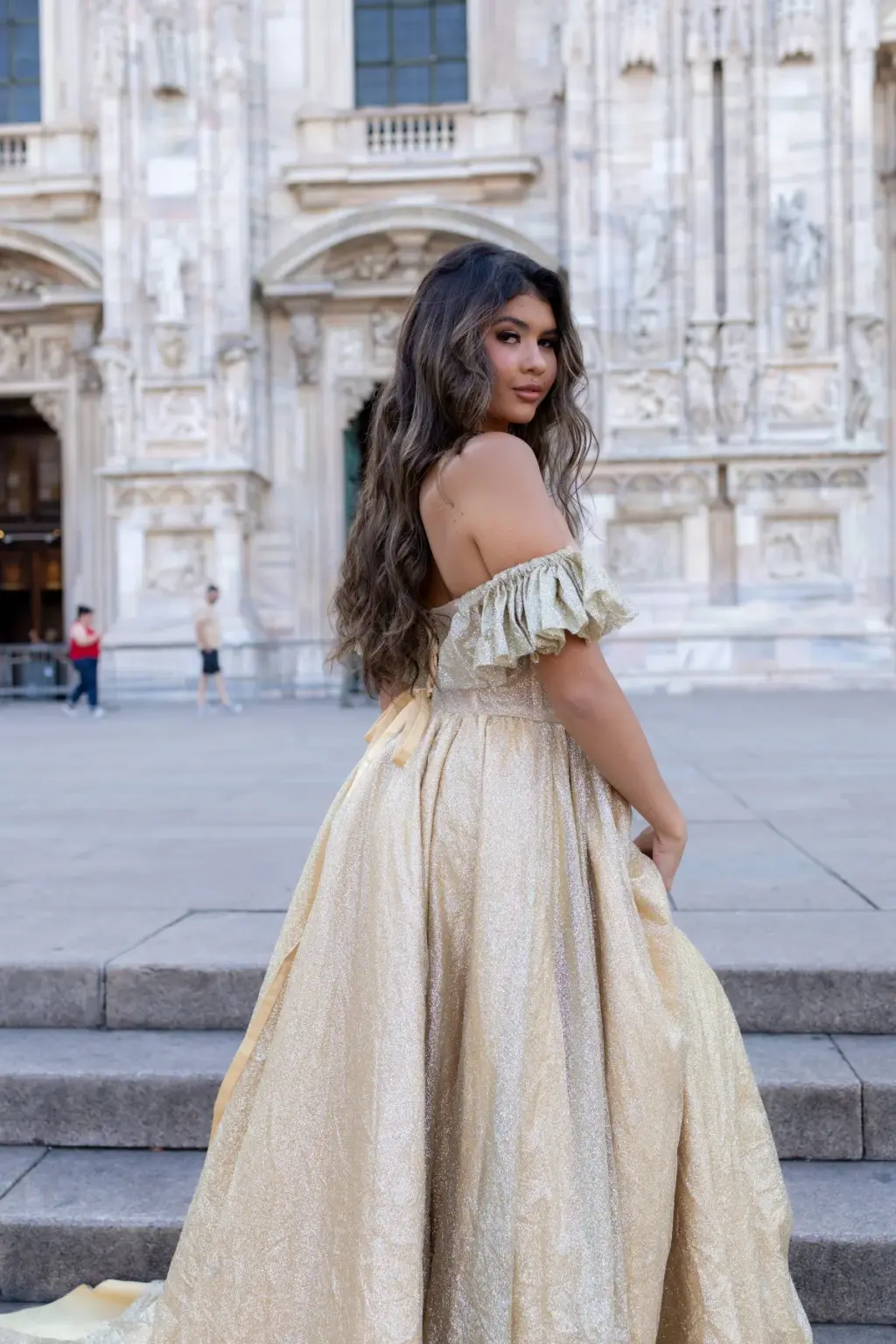 A woman in an off-the-shoulder gold gown climbs the Duomo steps and looks back over her shoulder at the camera.