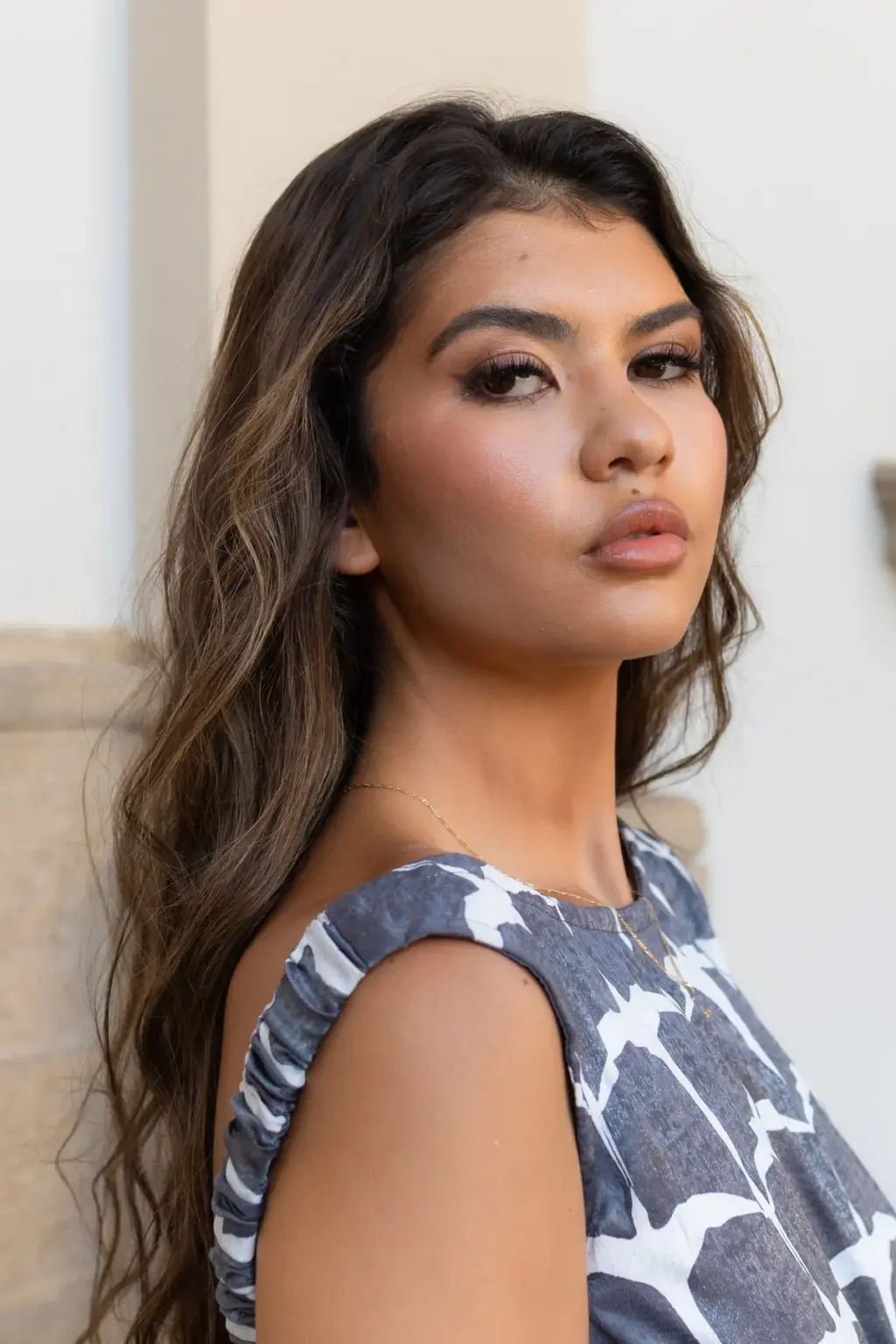 Tight portrait of a woman with glowing makeup and long wavy hair in a patterned top, turned toward the camera by a pale stone wall.