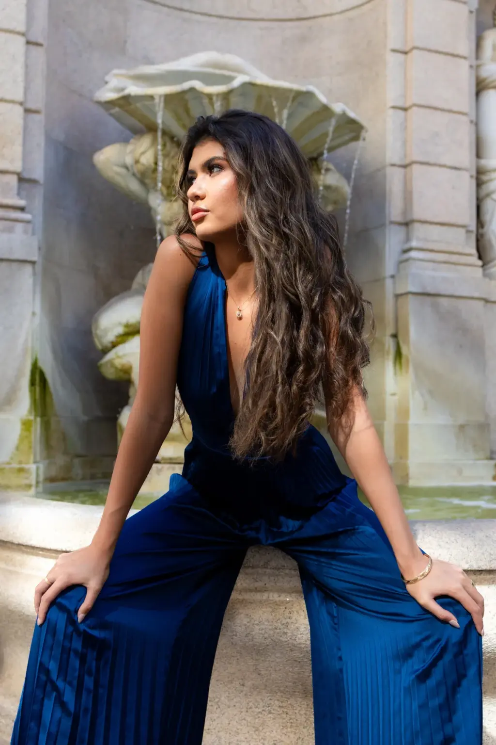 A woman with long dark hair sits back on a marble fountain edge in a navy pleated jumpsuit, gazing up and away from the camera.