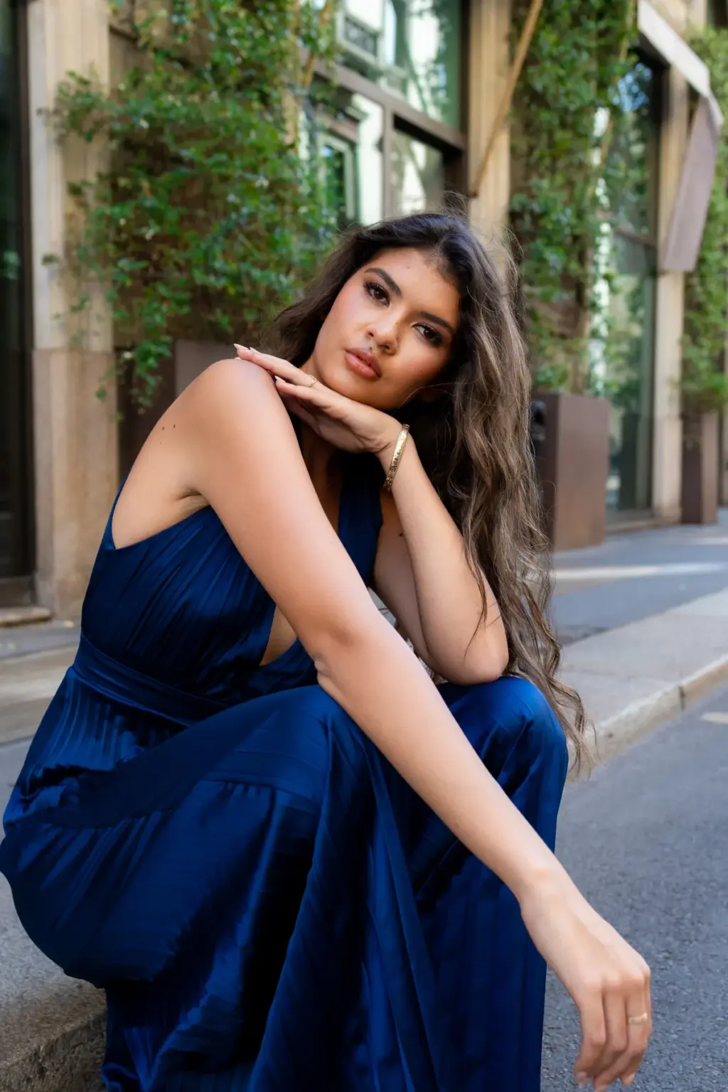 A woman in a navy pleated jumpsuit sits on a kerb, chin resting on her hand, against ivy-draped walls on a Milan side street.