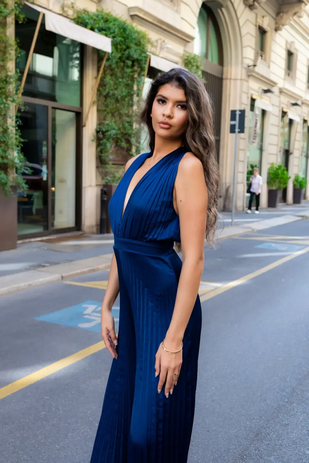 A woman in a pleated navy halter jumpsuit stands in the middle of a quiet Milan side street lined with ivy-covered facades.