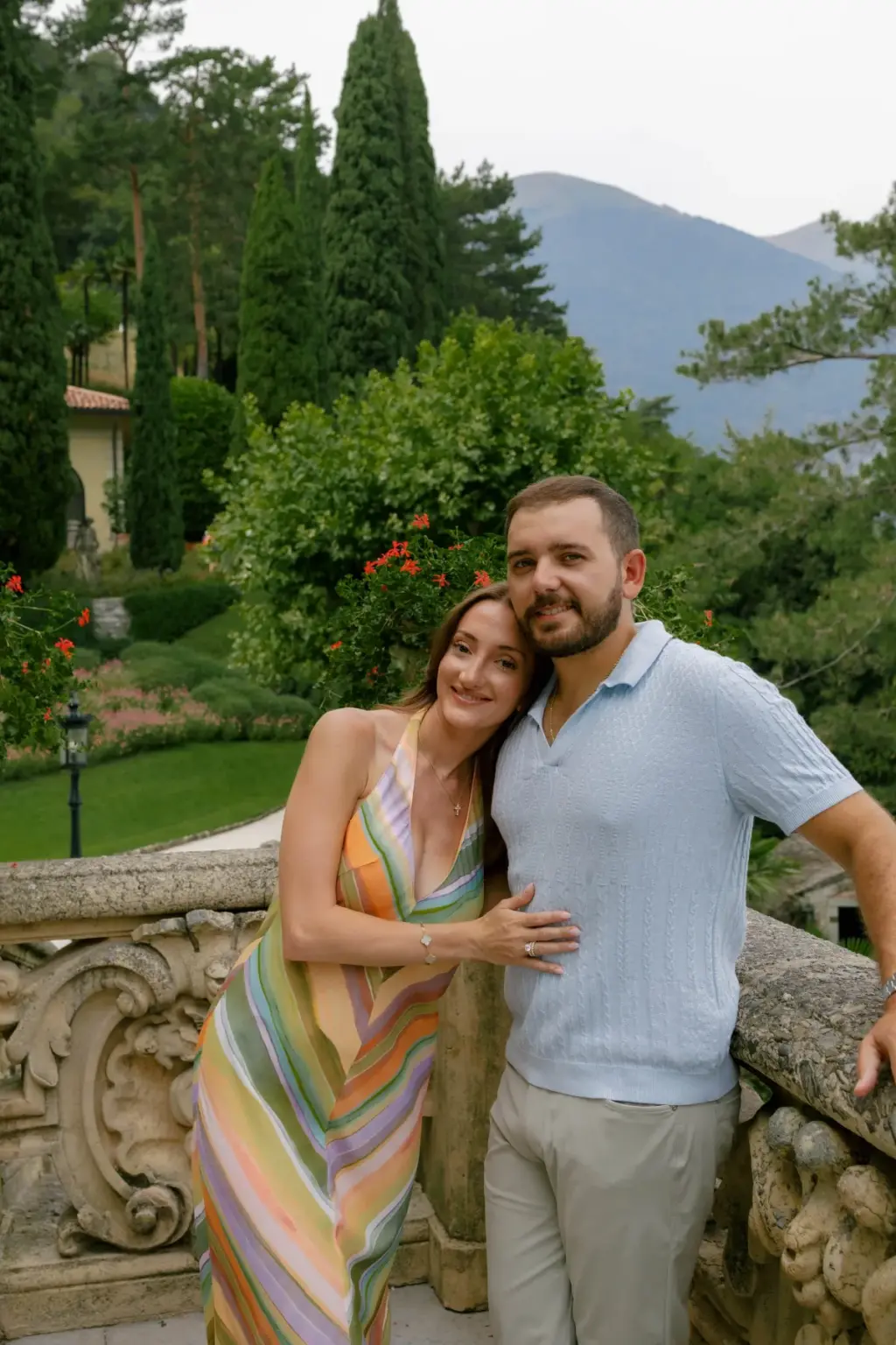 A smiling couple leans on a carved balustrade at Villa del Balbianello, cypress trees, red flowers and mountains behind them.
