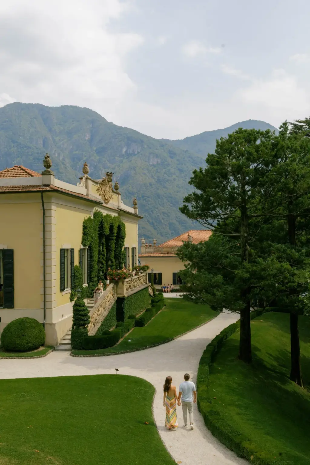 Wide view of a couple walking hand in hand along a curving garden path at Villa del Balbianello, the villa and green hills beside them.