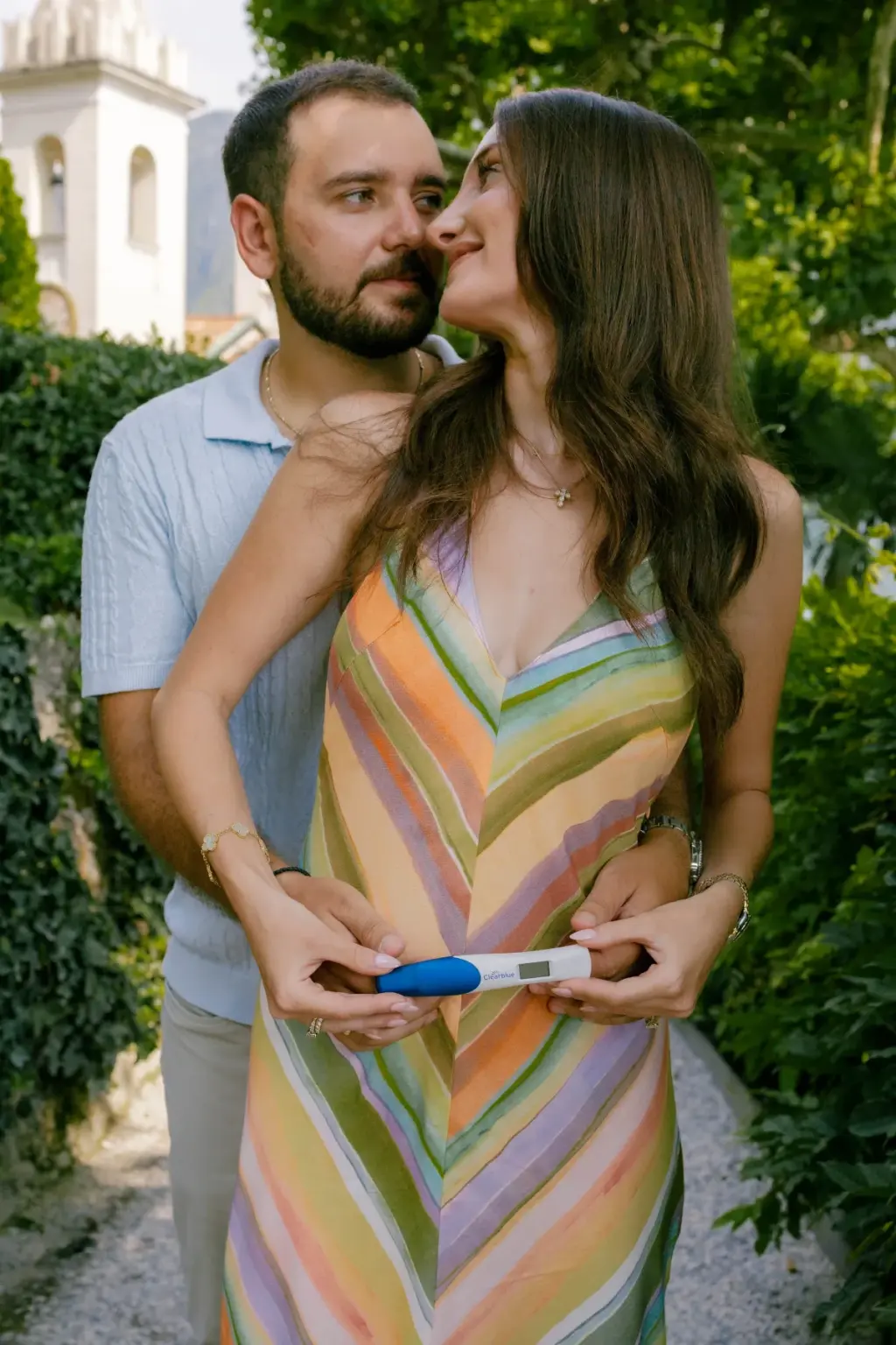 A man hugs his partner from behind and kisses her cheek as she holds a pregnancy test at her waist on a green garden path.