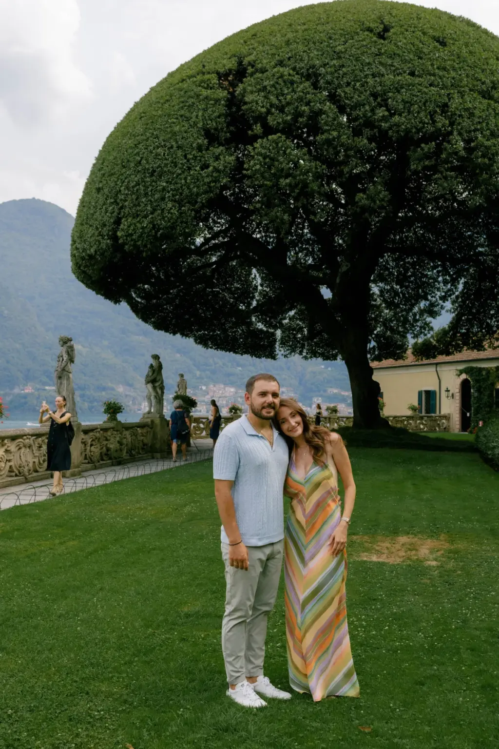 A couple stands arm in arm on the lawn under the great umbrella-shaped tree at Villa del Balbianello, statues along the wall.