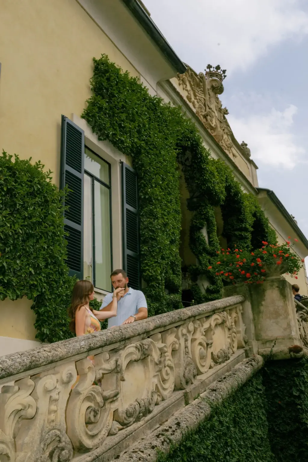 A couple shares a quiet moment on the carved stone balcony of Villa del Balbianello, the wall draped in ivy and red geraniums.