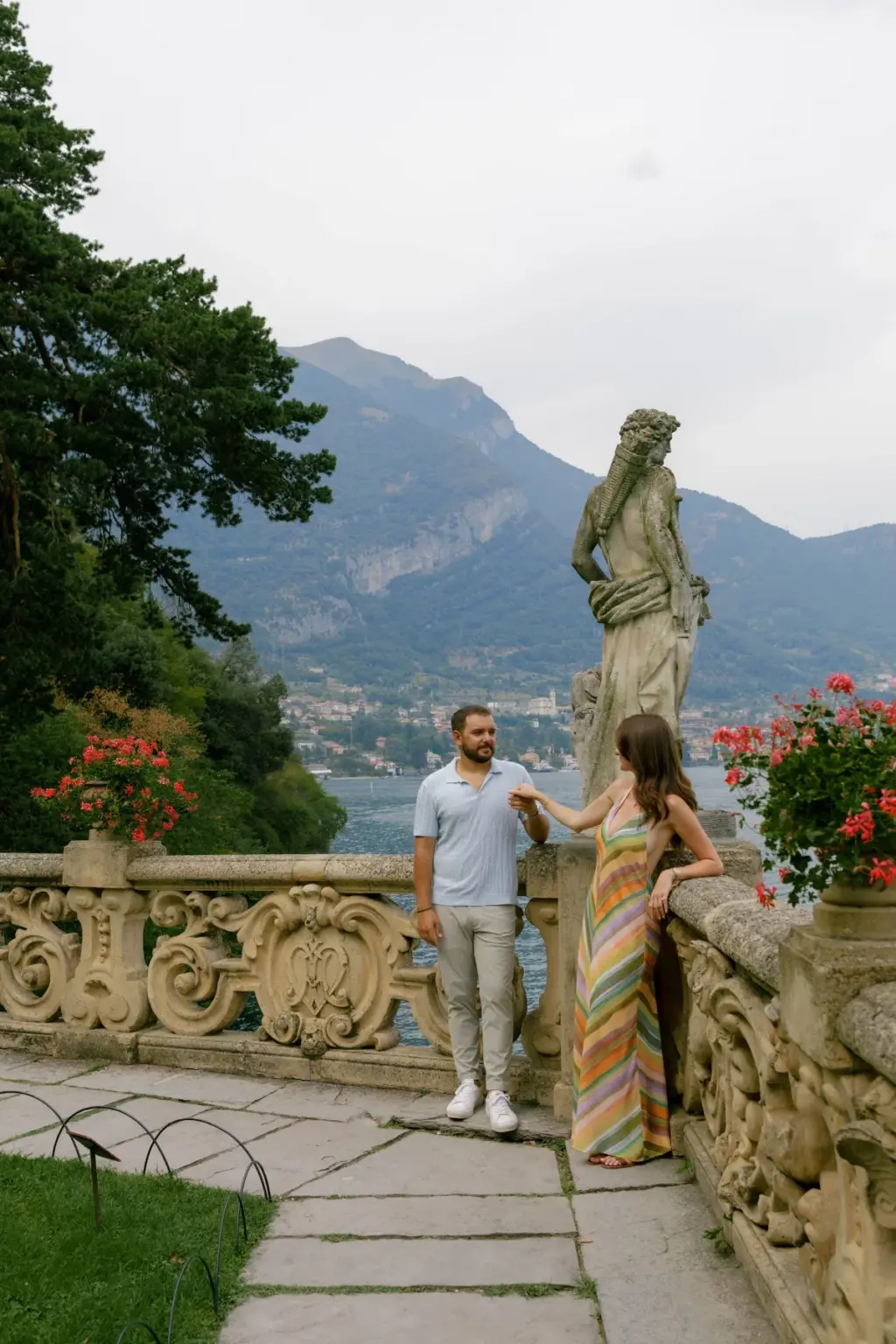 A woman takes her partner's hand by a stone statue on the Villa del Balbianello terrace, with Lake Como and mountains beyond.