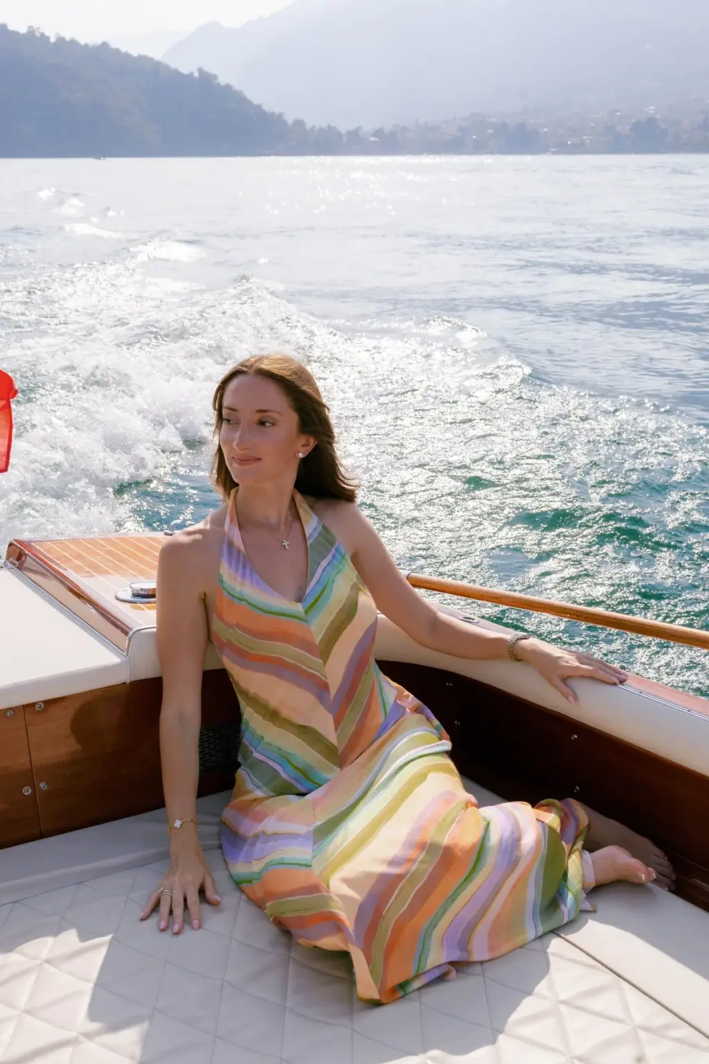 A woman in a rainbow-striped dress sits at the stern of a wooden boat, the white wake trailing across Lake Como behind her.