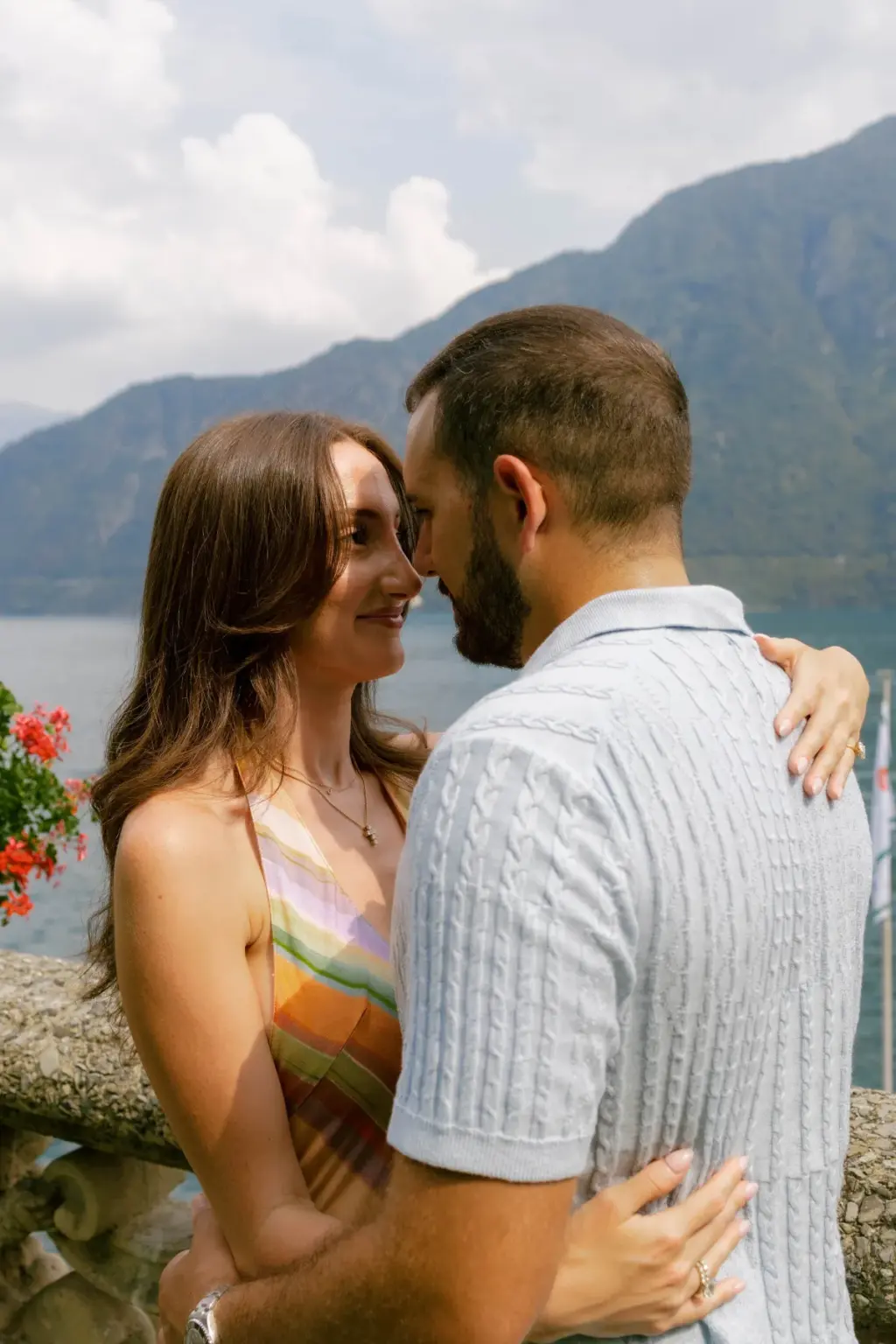 A bearded man in a knitted polo holds his partner close as they smile into each other's eyes against a Lake Como mountain view.
