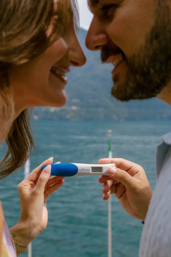 Close-up of a laughing couple nose to nose, holding a pregnancy test between them above the blue water of Lake Como.
