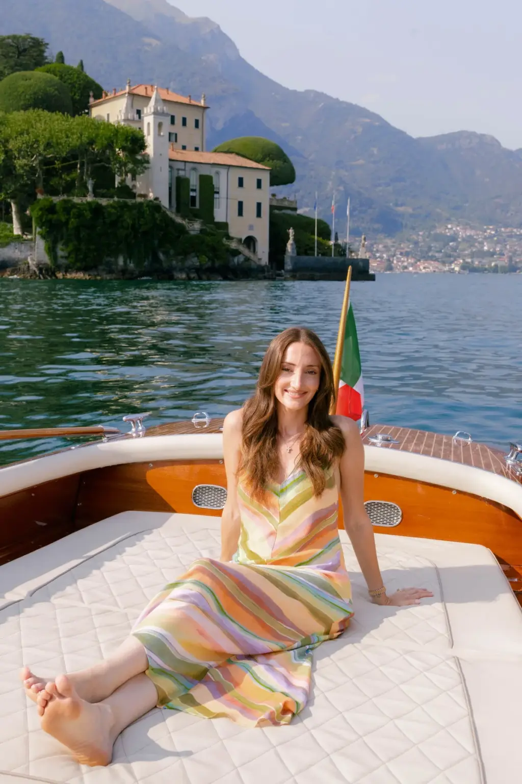 A woman in a pastel striped maxi dress sits barefoot on the cushions of a wooden boat, Villa del Balbianello across the water.