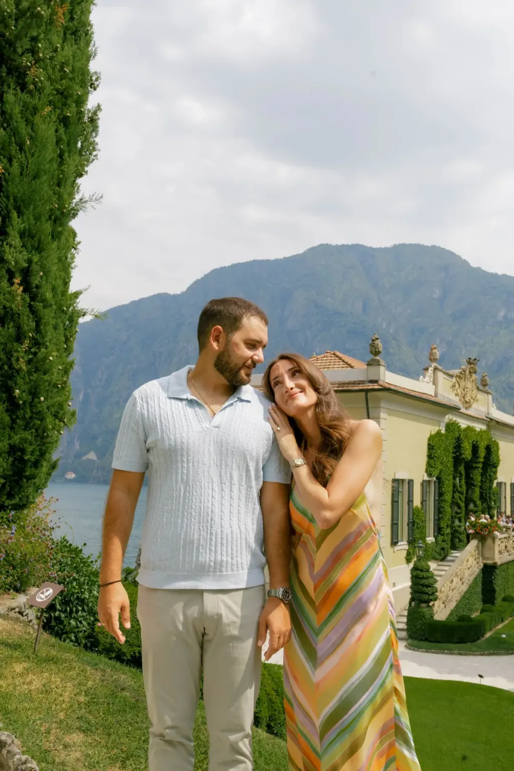 A woman in a striped pastel maxi dress leans her head toward her bearded partner on a lawn at Villa del Balbianello, mountains behind.