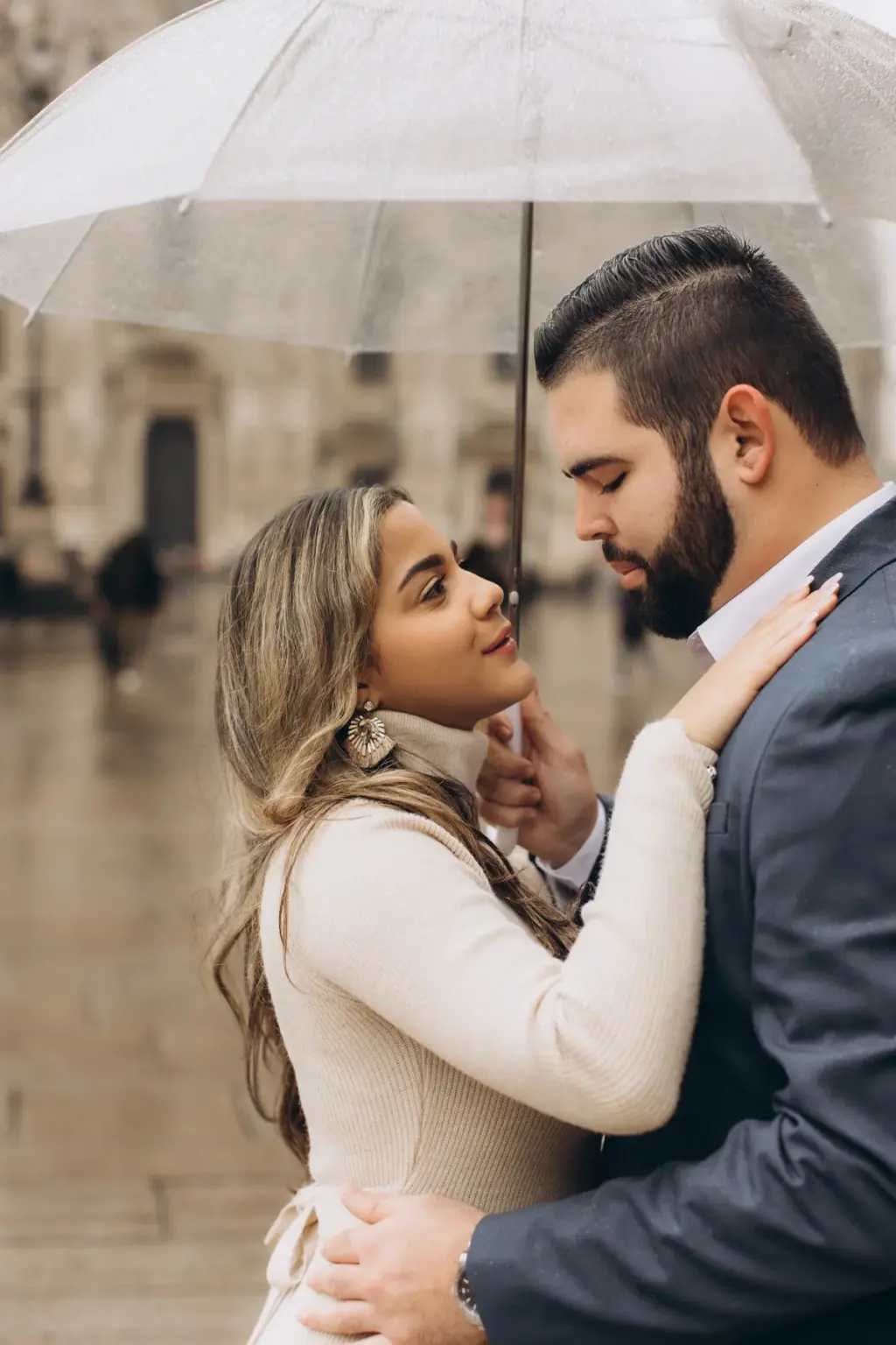 A couple shares a close moment under a clear umbrella on the rain-wet Duomo square, the woman's hand resting on his collar.