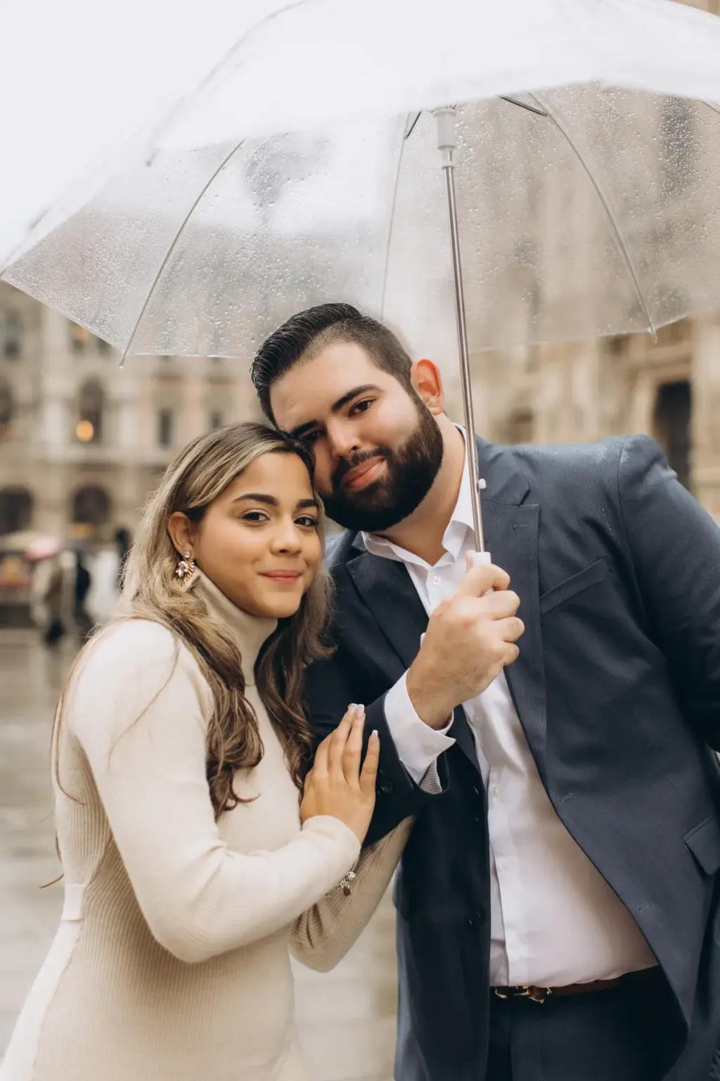 A couple leans head to head beneath a clear umbrella in the rain, the woman's ringed hand on his chest, the Duomo blurred behind.