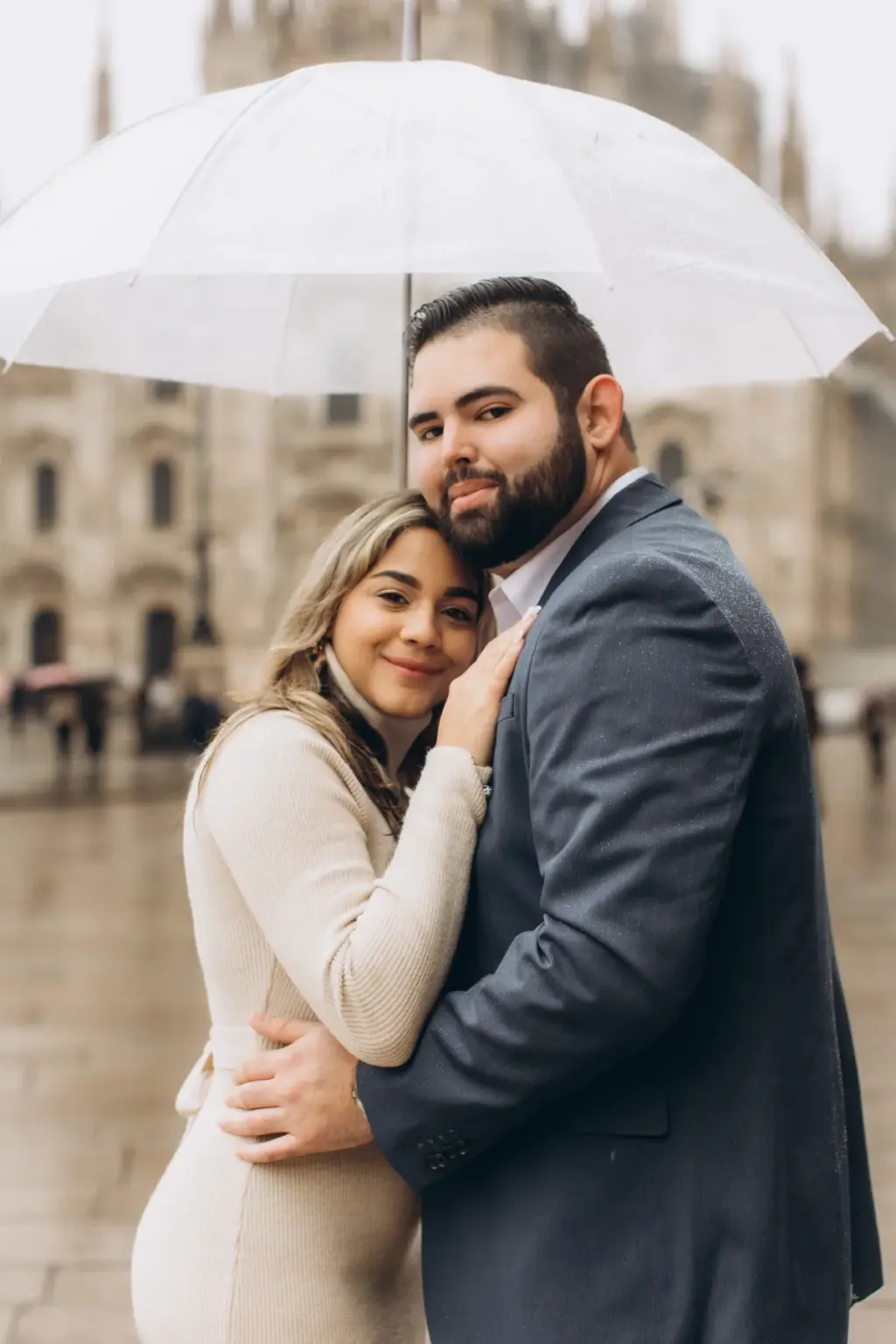 A woman leans her head on her partner's chest under a clear umbrella, the Duomo facade misty behind them on a rainy day.