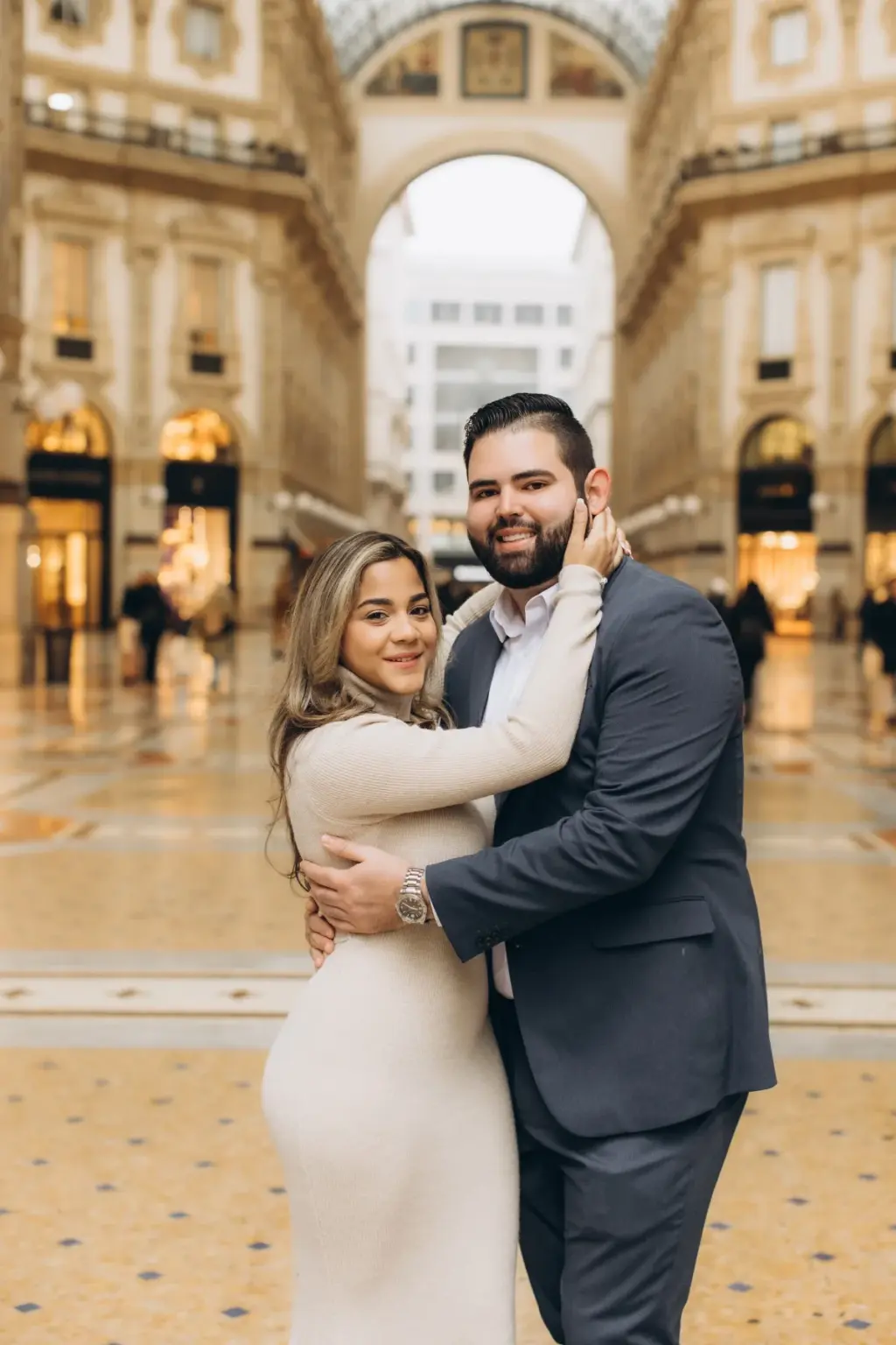 A woman wraps her arms around her partner's neck and smiles at the camera on the patterned floor of the Galleria under its arch.