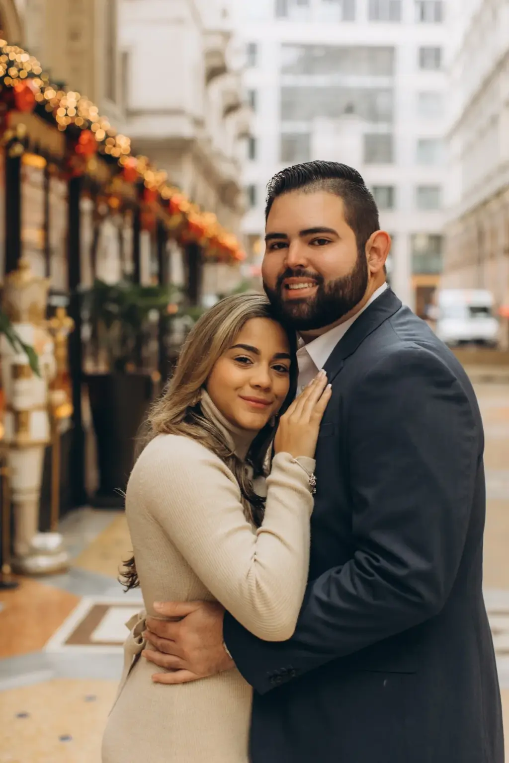 A smiling couple hugs cheek to shoulder, the woman's hand on his chest, beneath glowing Christmas garlands in the Galleria.