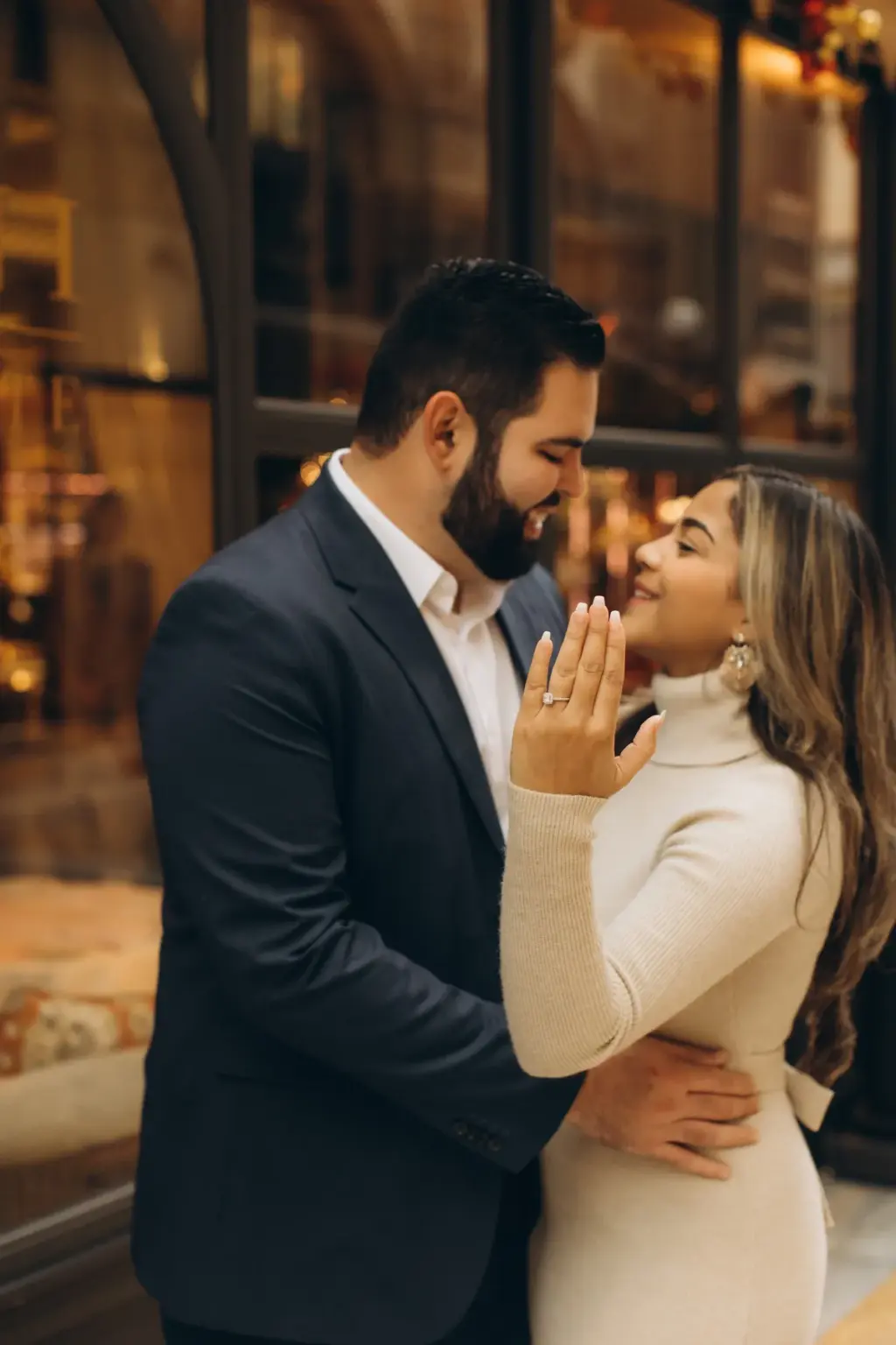 A woman in a cream turtleneck holds up her hand to show an engagement ring as her bearded partner draws her close in the Galleria.