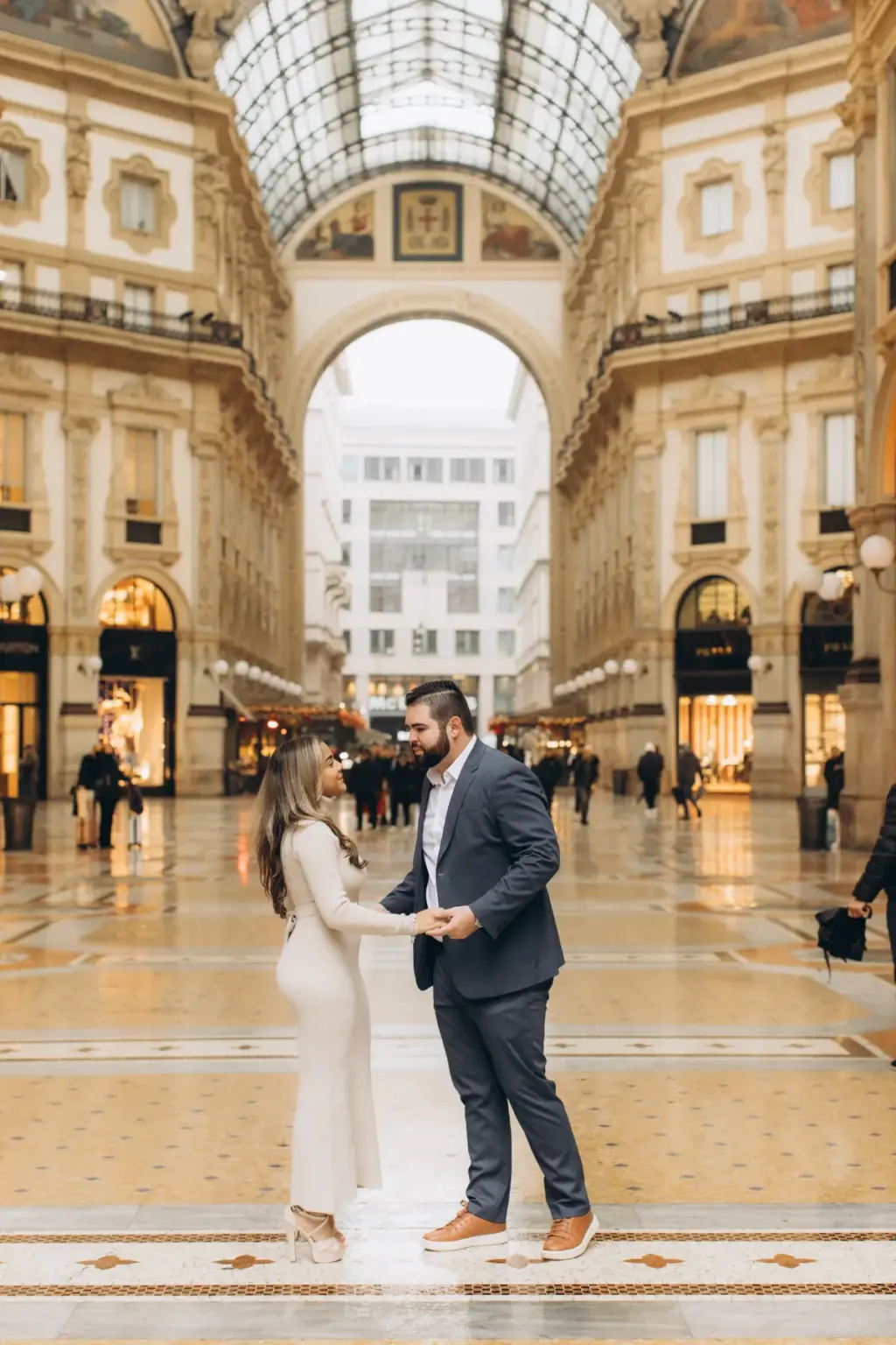 A couple holds hands facing each other in the middle of the Galleria Vittorio Emanuele II, the glass roof and grand arch behind.