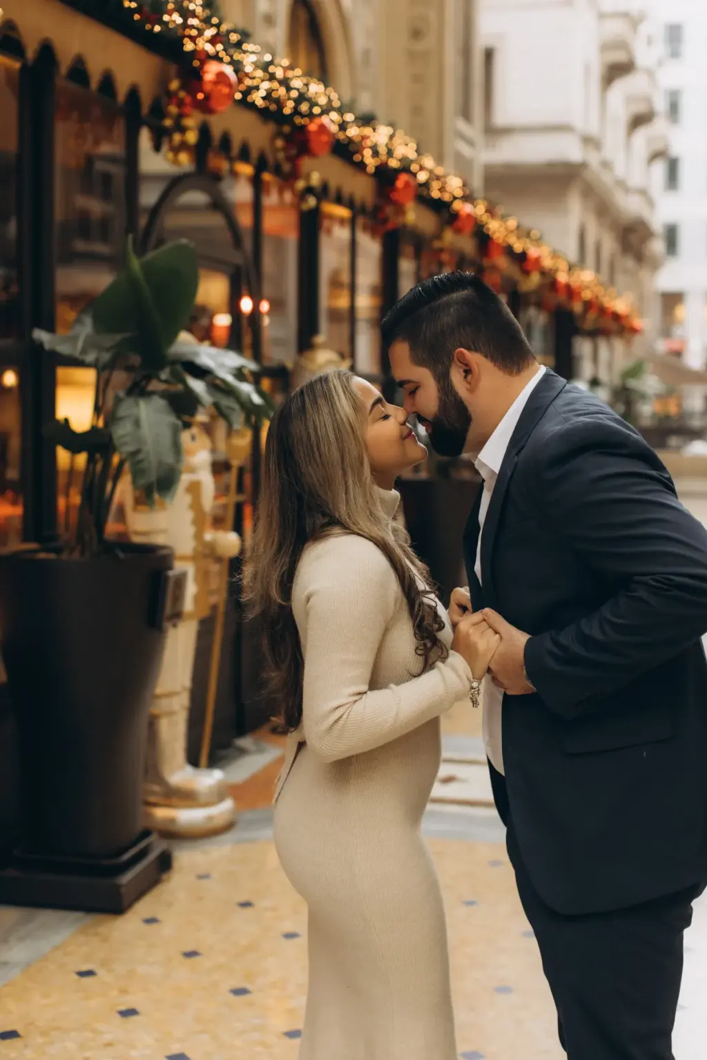 A bearded man in a navy suit and a woman in a cream knit dress lean in for a kiss under Christmas garlands outside Savini in the Galleria.