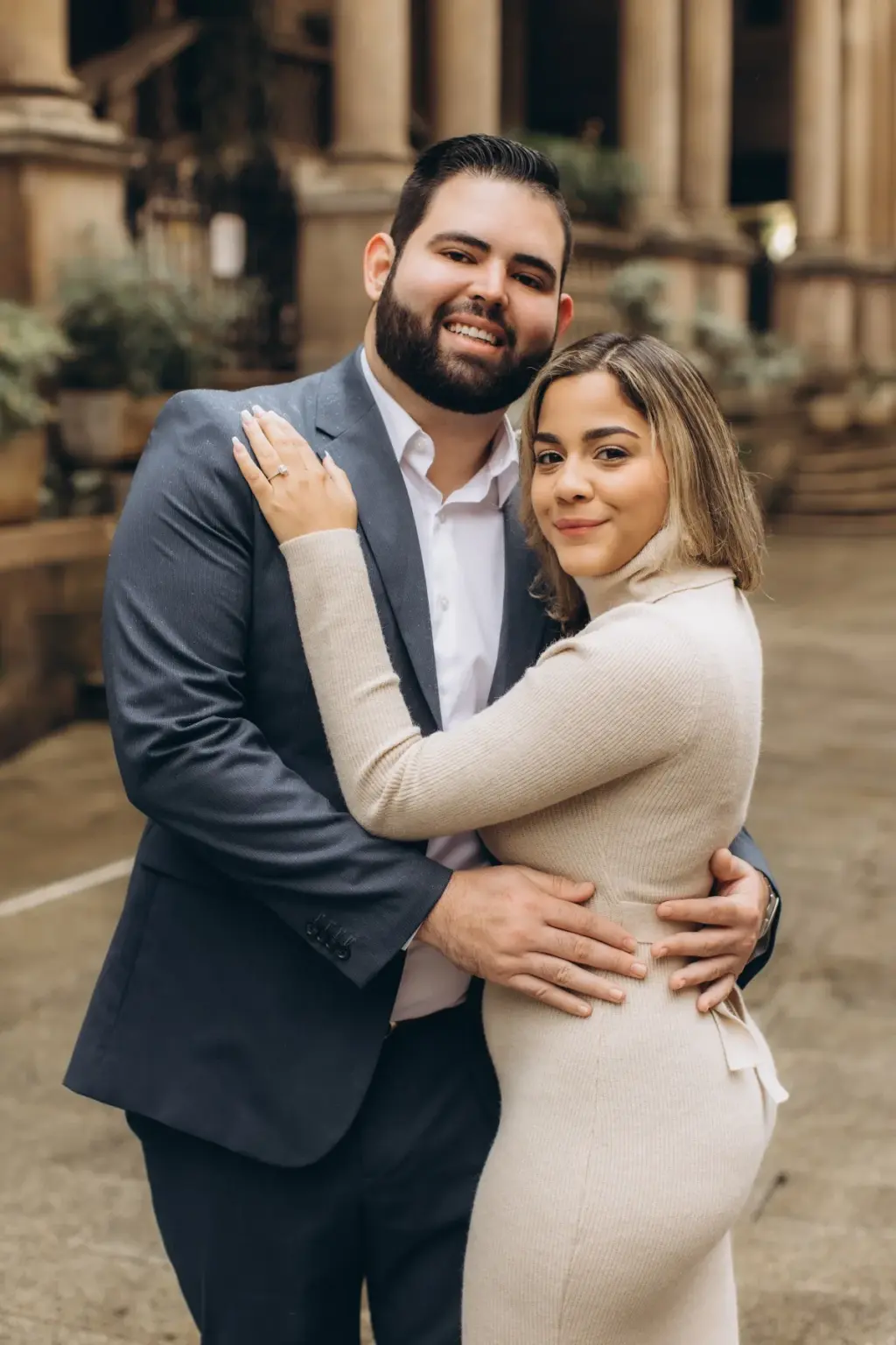 A smiling couple in a tight embrace pose by the stone columns of an arcade near Piazza dei Mercanti, her hand on his shoulder.