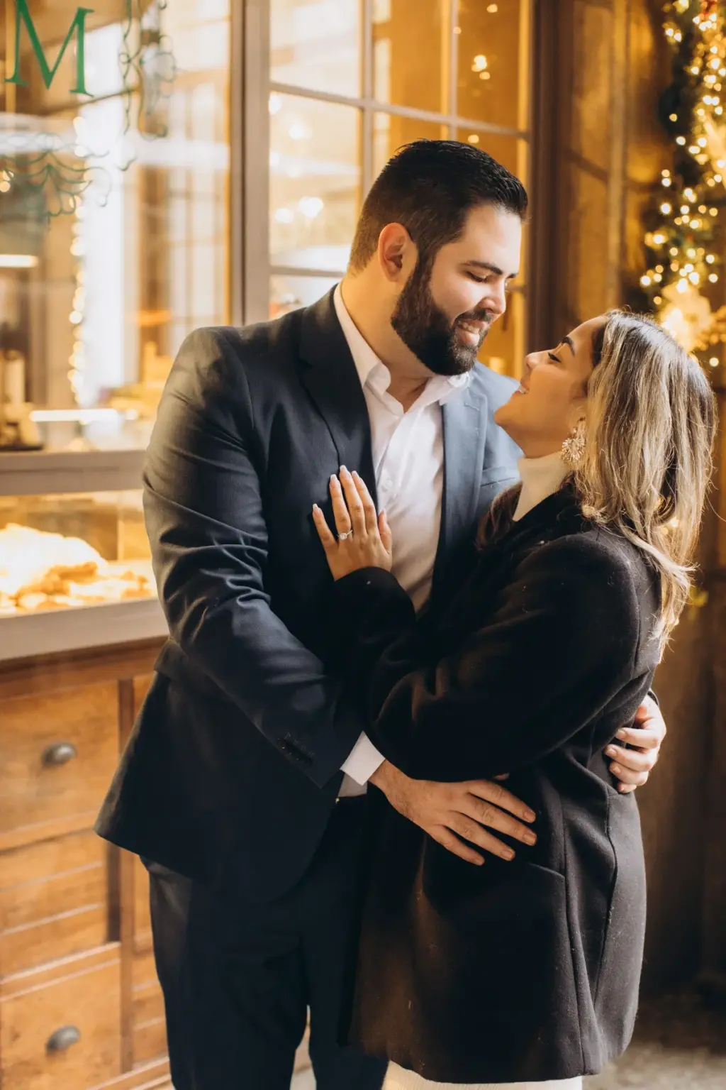 A woman in a black coat laughs up at her partner, ring on her hand at his chest, in front of a bakery window strung with fairy lights.
