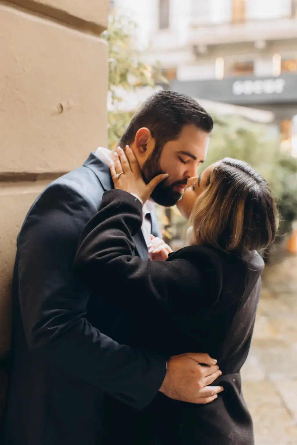 A woman in a dark coat pulls her bearded partner in for a kiss as he leans against a pale stone wall on a Milan side street.