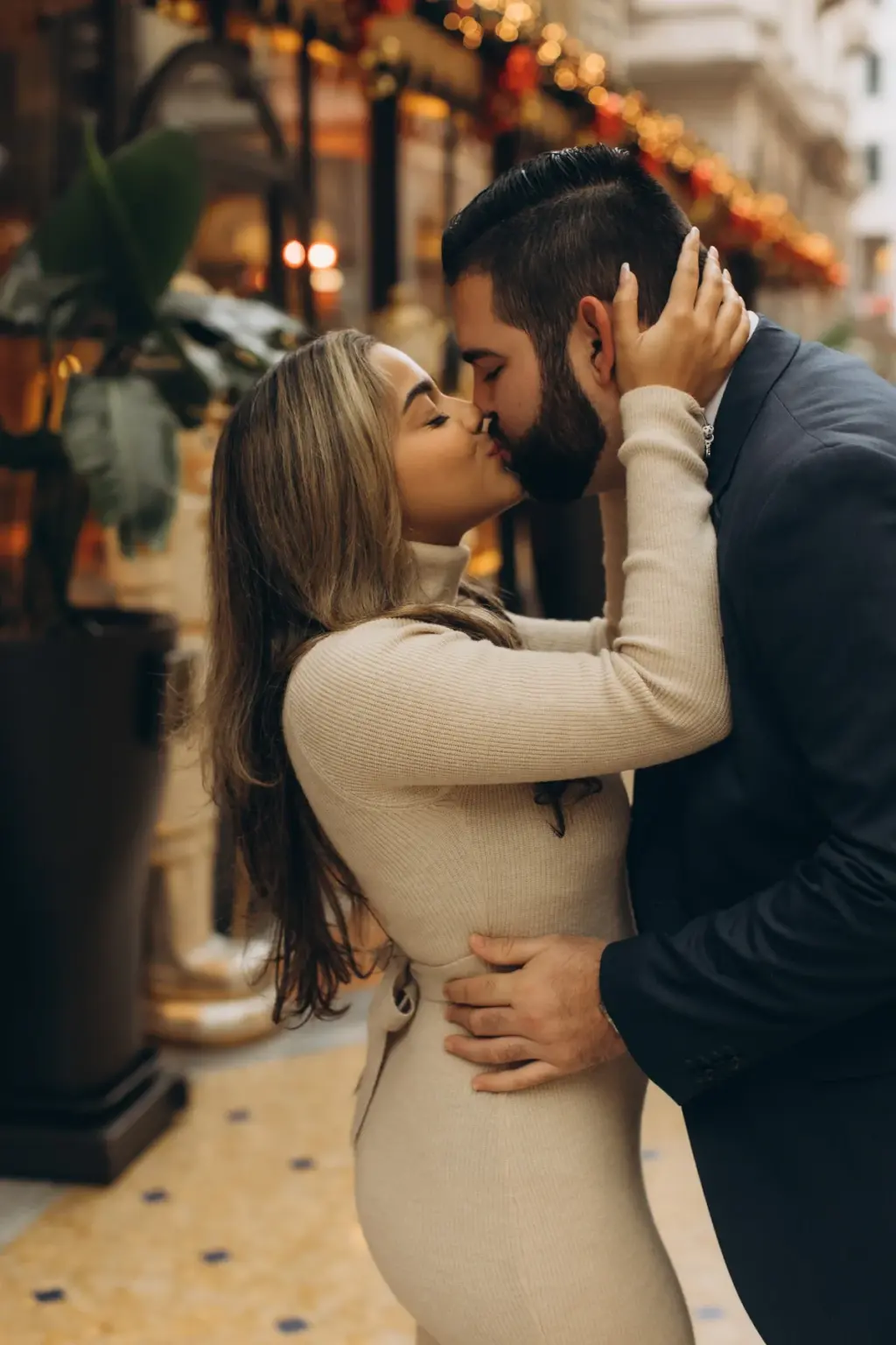 A woman in a cream ribbed dress cups her partner's face as they kiss, his hand at her waist, by the festive Savini entrance.