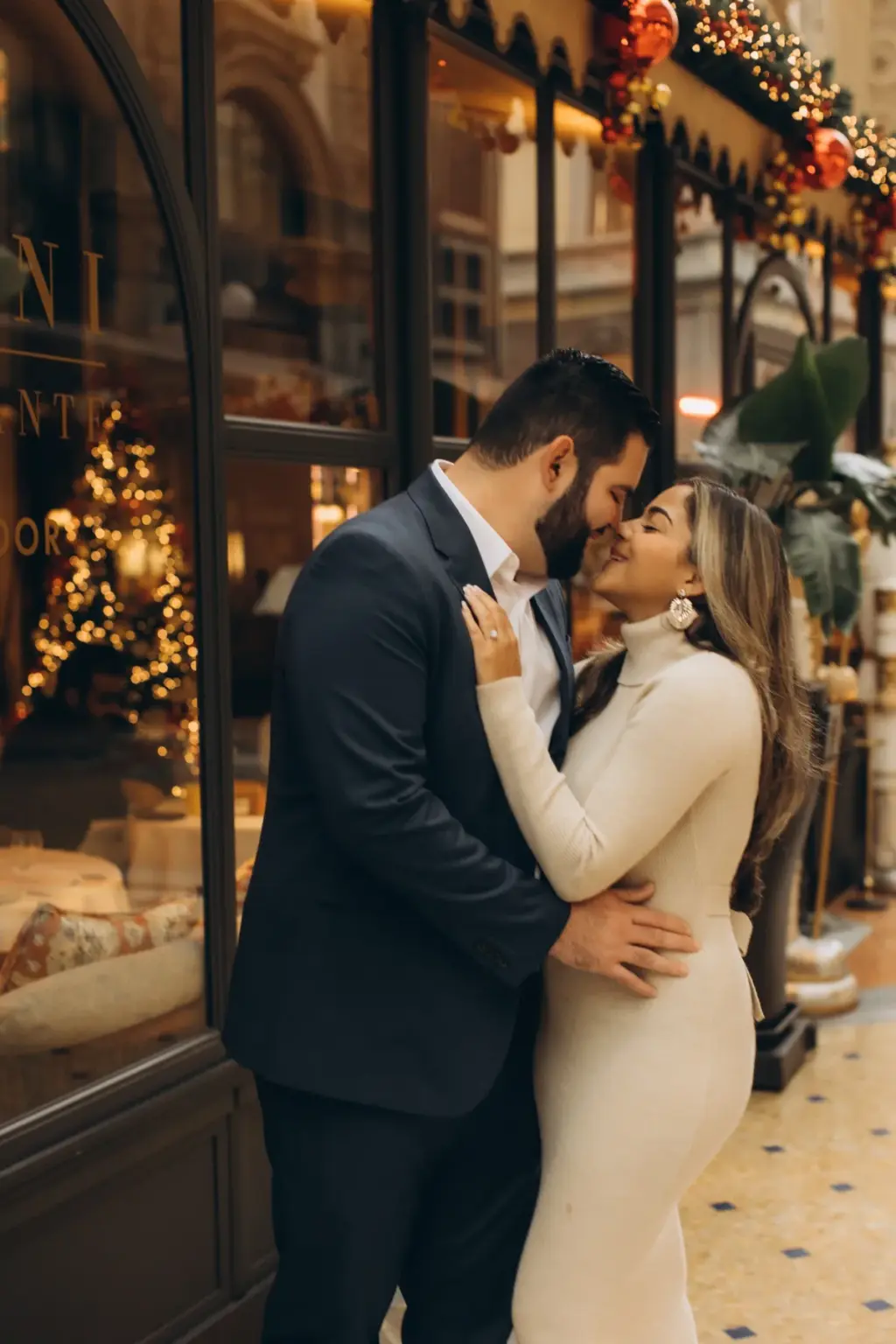 A man presses his face to his partner's cheek as she smiles with eyes closed, framed by Savini's festive shopfront in Milan.