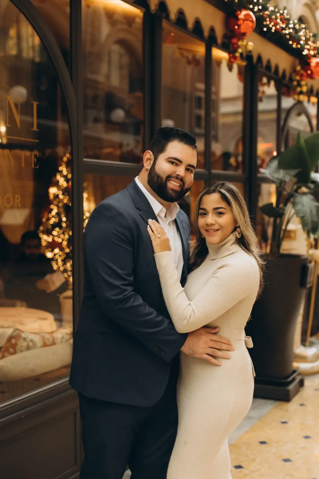 A happy couple stands embraced in front of the Savini window with its little Christmas tree, her ringed hand on his chest.