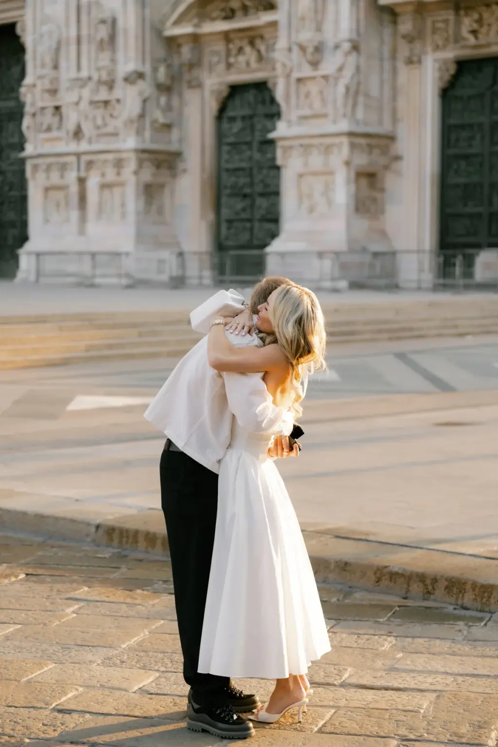 A woman in a white midi dress throws her arms around her partner in a cream jacket on the square in front of the Duomo doors.