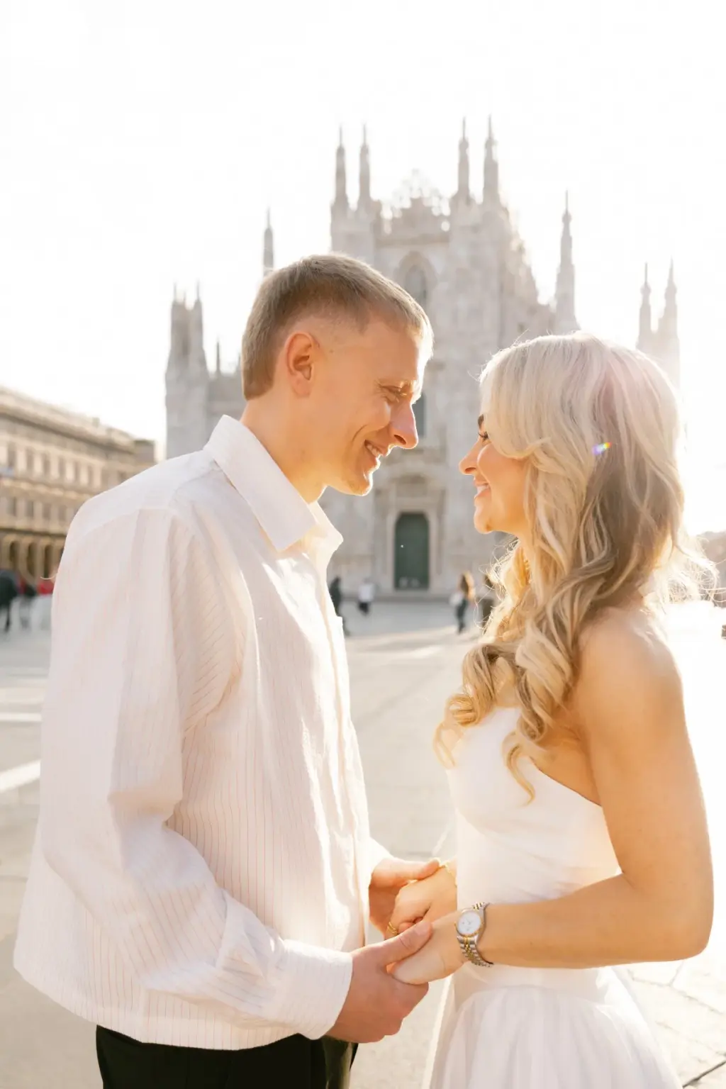 Smiling, a couple hold both hands and lean toward each other with the Duomo spires softly blurred behind them in bright light.