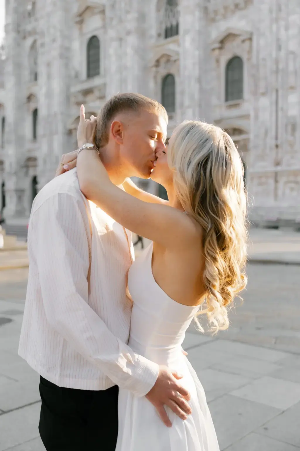 A man in a white shirt kisses a woman in a white strapless dress as she wraps her arm around his neck in front of the Duomo facade.