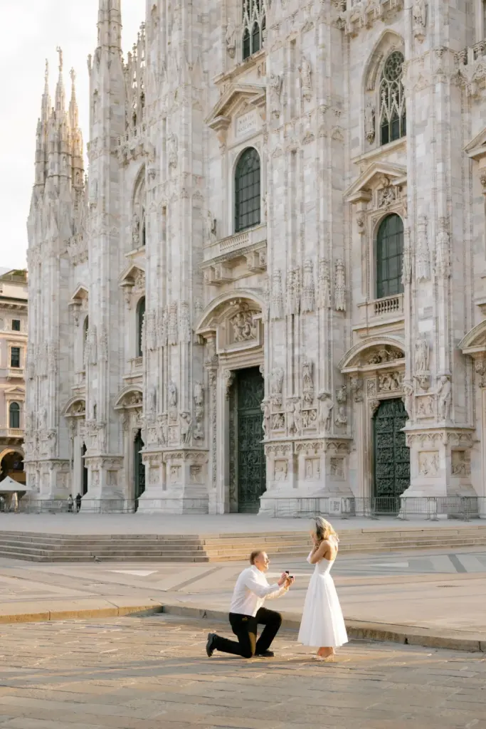 A man kneels on the empty Piazza del Duomo to propose to a woman in a white dress, the cathedral facade rising behind them.