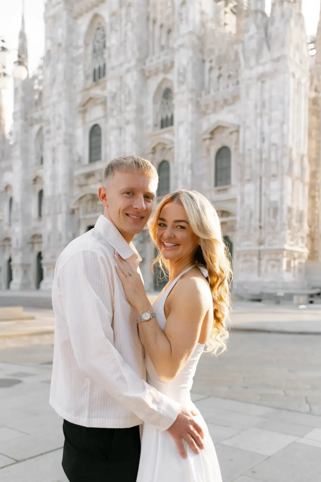 A couple smile at the camera in front of the Duomo, her ringed hand resting on his chest and his arm wrapped around her waist.