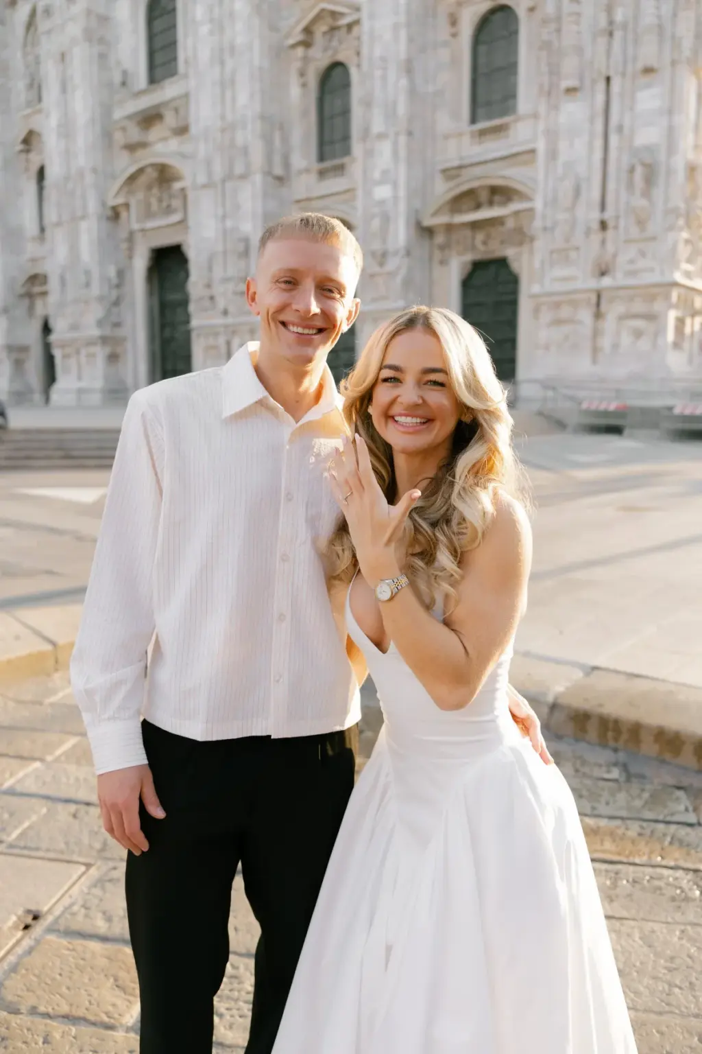 A smiling couple stands before the Duomo di Milano, the blonde woman in white holding up her hand to show her new ring.