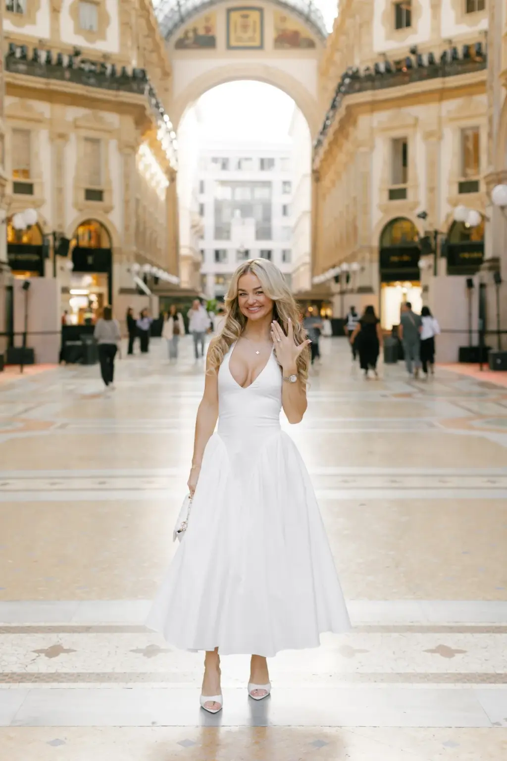 Under the great arch of the Galleria, a blonde woman in a white dress lifts her hand to show her ring and smiles at the camera.