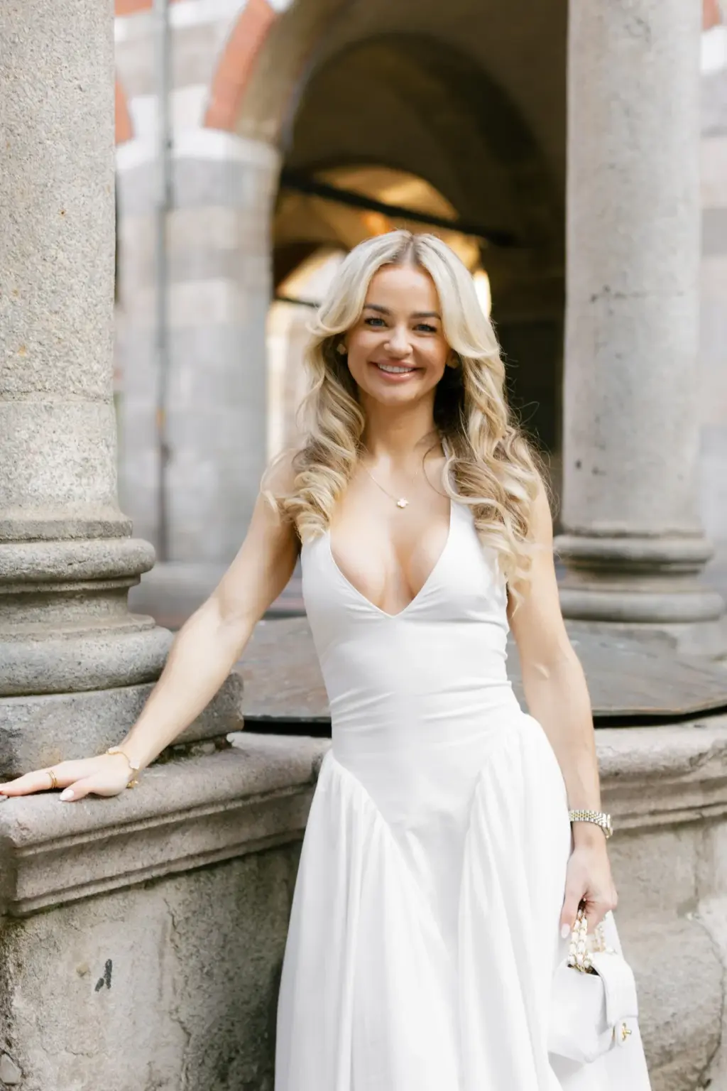 A smiling blonde woman in a white dress leans against a stone column under the arches of the Loggia dei Mercanti.