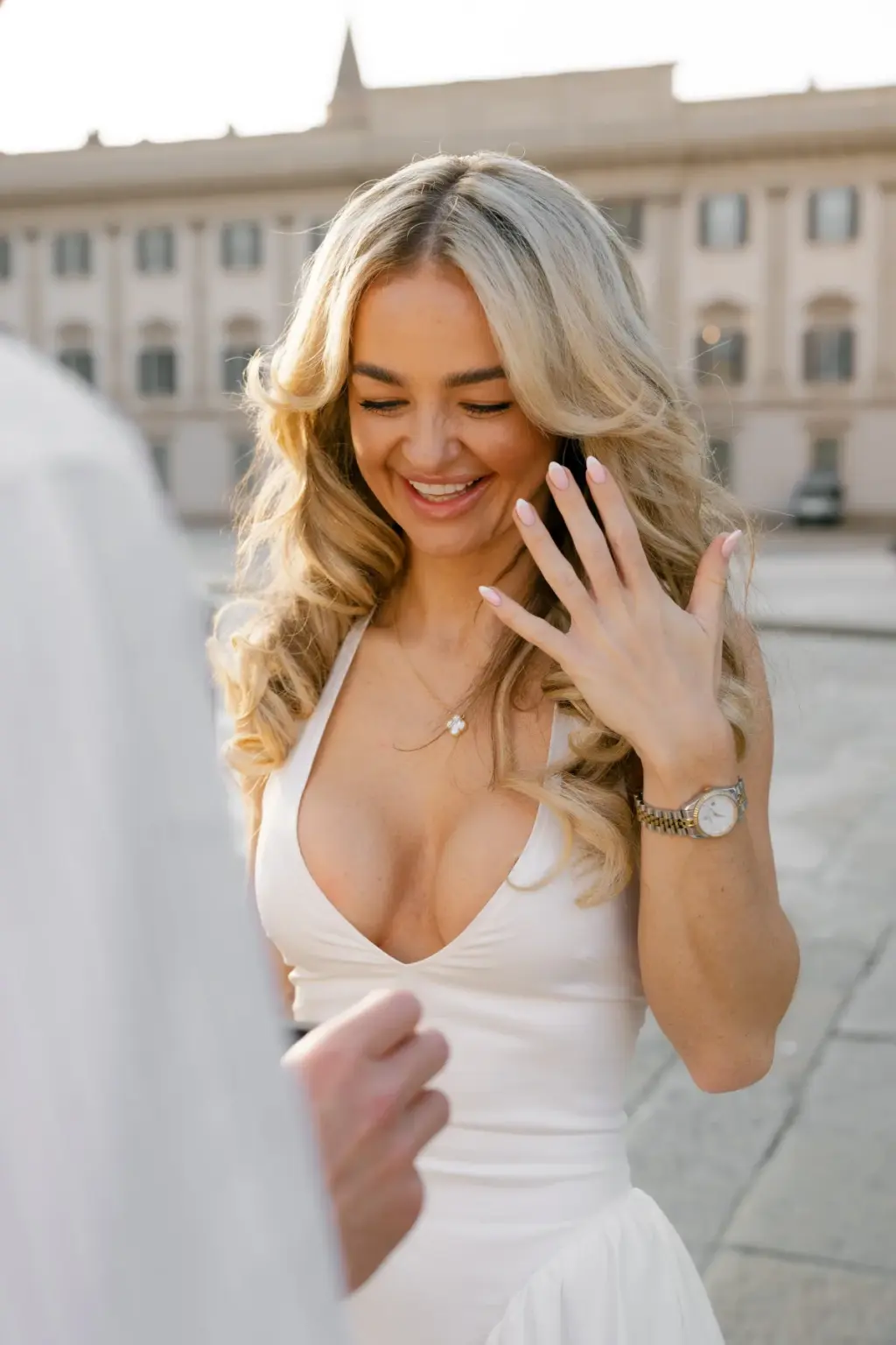 A blonde woman in a white dress laughs with eyes nearly shut as she looks down at the ring on her hand in Piazza del Duomo.