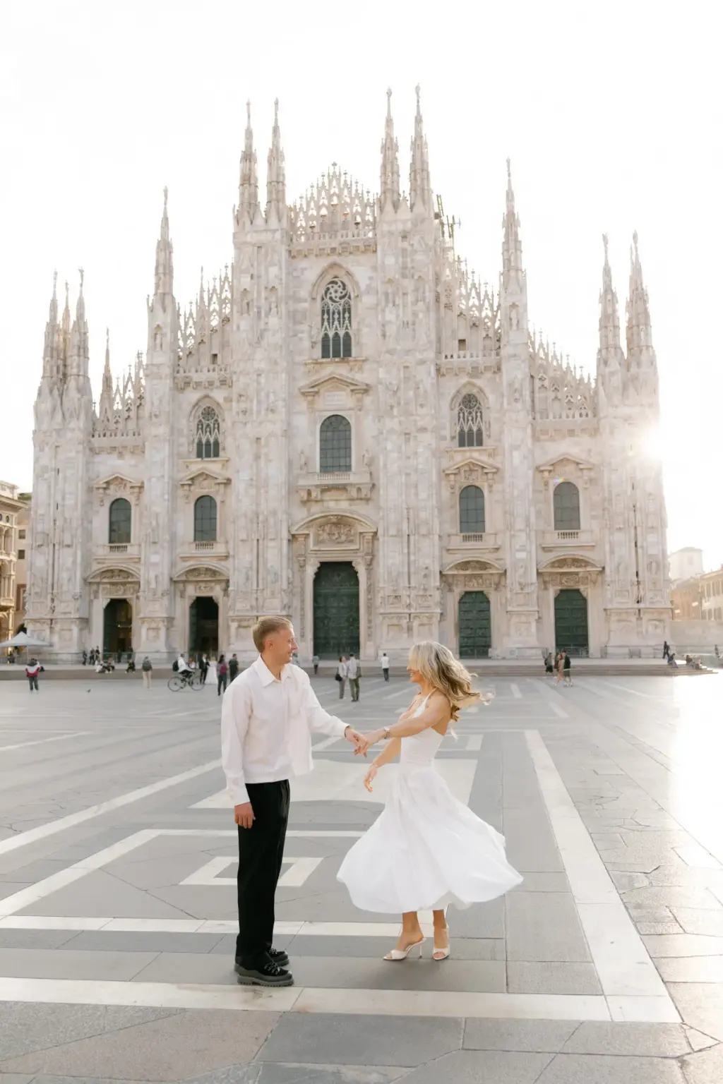 Hand in hand, a man spins a woman in a flowing white dress across the wide open Piazza del Duomo with the cathedral behind them.
