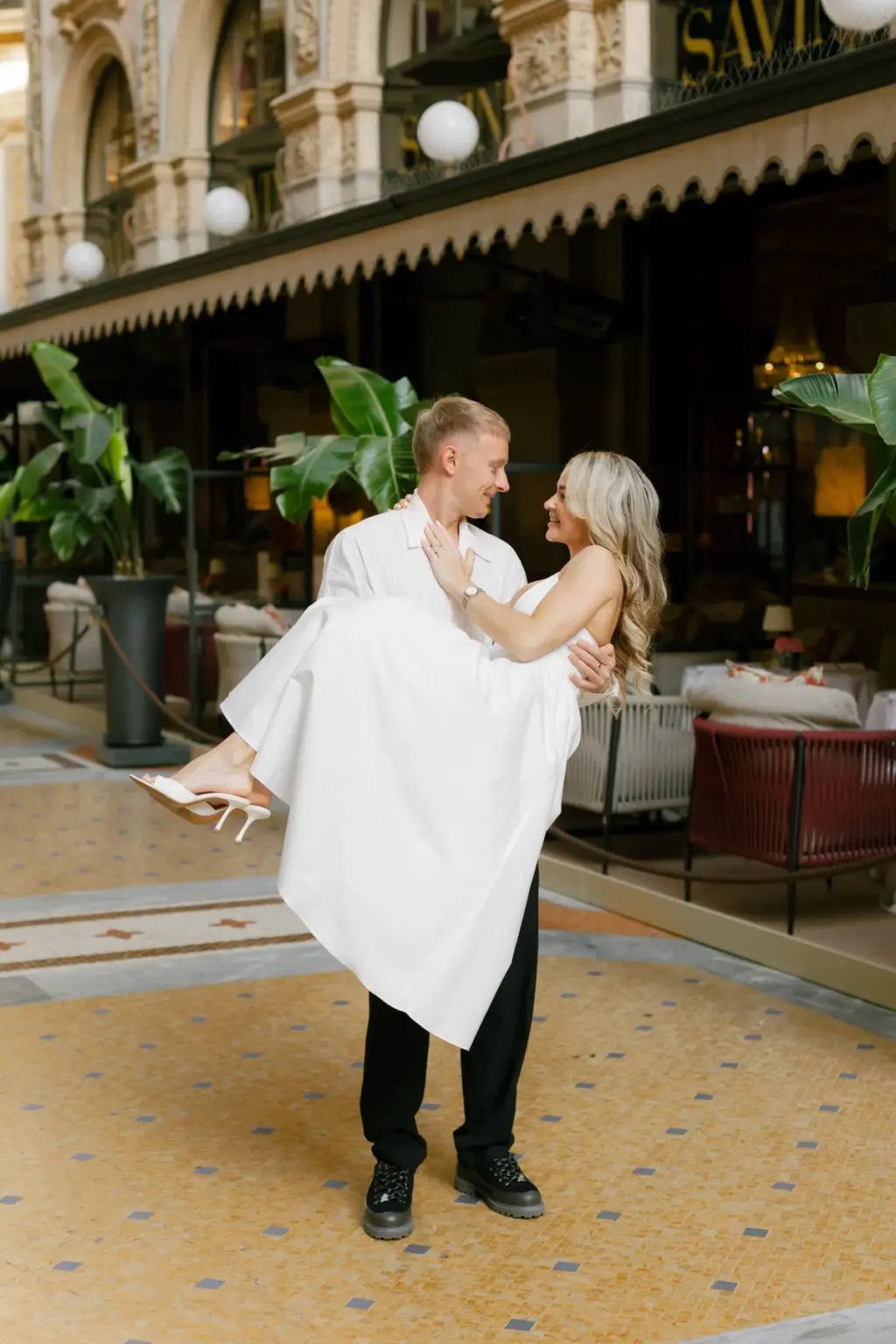 A man carries a woman in a white dress in his arms past the green-and-gold awning of Savini inside the Galleria.