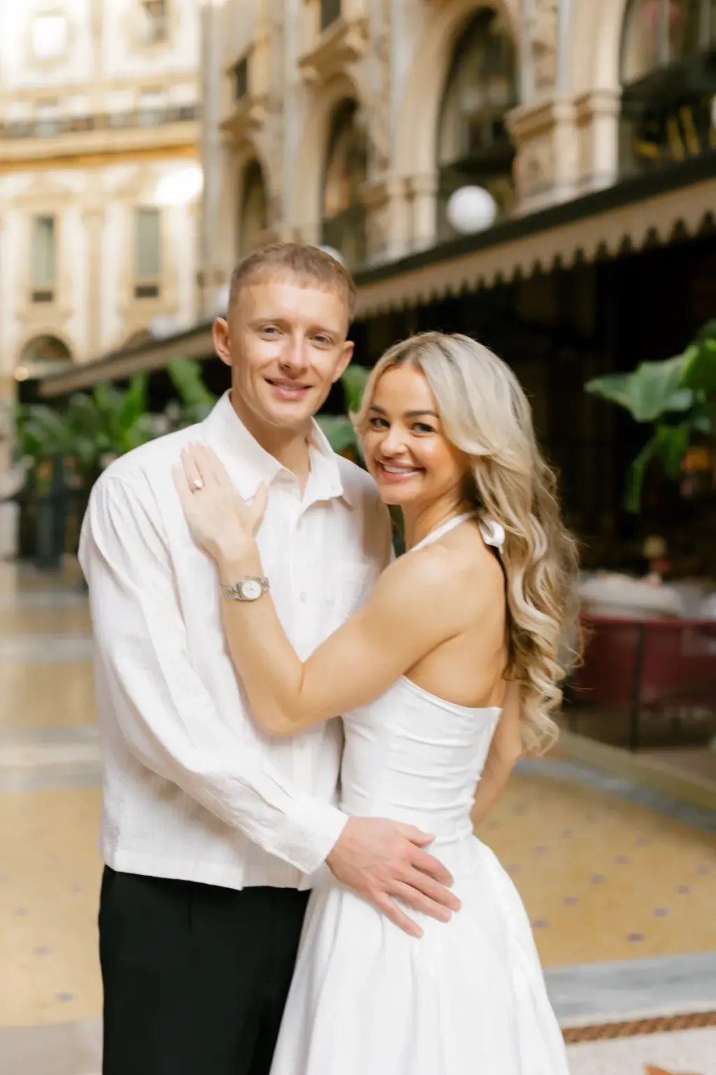 A couple poses arm in arm in front of the Savini restaurant terrace in the Galleria, both smiling straight at the camera.
