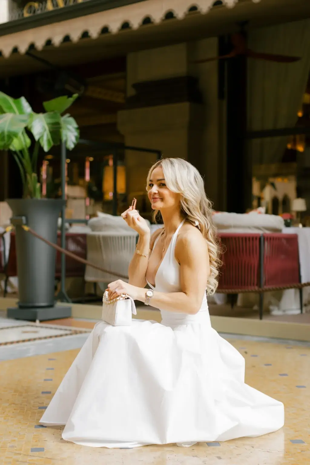 A woman in a white dress crouches on the mosaic floor by the Savini terrace, gazing at her ring with a small white bag in her lap.